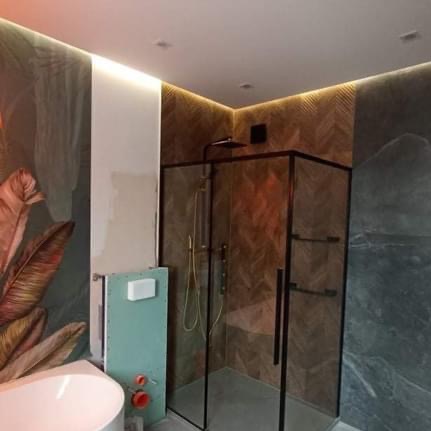 Nowoczesna łazienka w trakcie wykańczania: kabina prysznicowa z czarnymi profilami, ściana z motywem liści, podświetlane krawędzie sufitu, zabudowa podtynkowa toalety w kolorze miętowym.