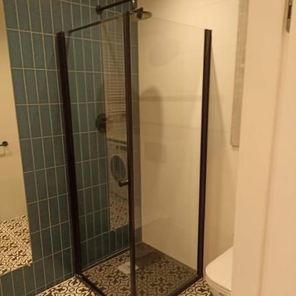 Nowoczesna kabina prysznicowa z czarnymi profilami i szklanymi ściankami, widoczna na tle turkusowych płytek ułożonych pionowo oraz wzorzystej podłogi. W tle pralka i fragment toalety.