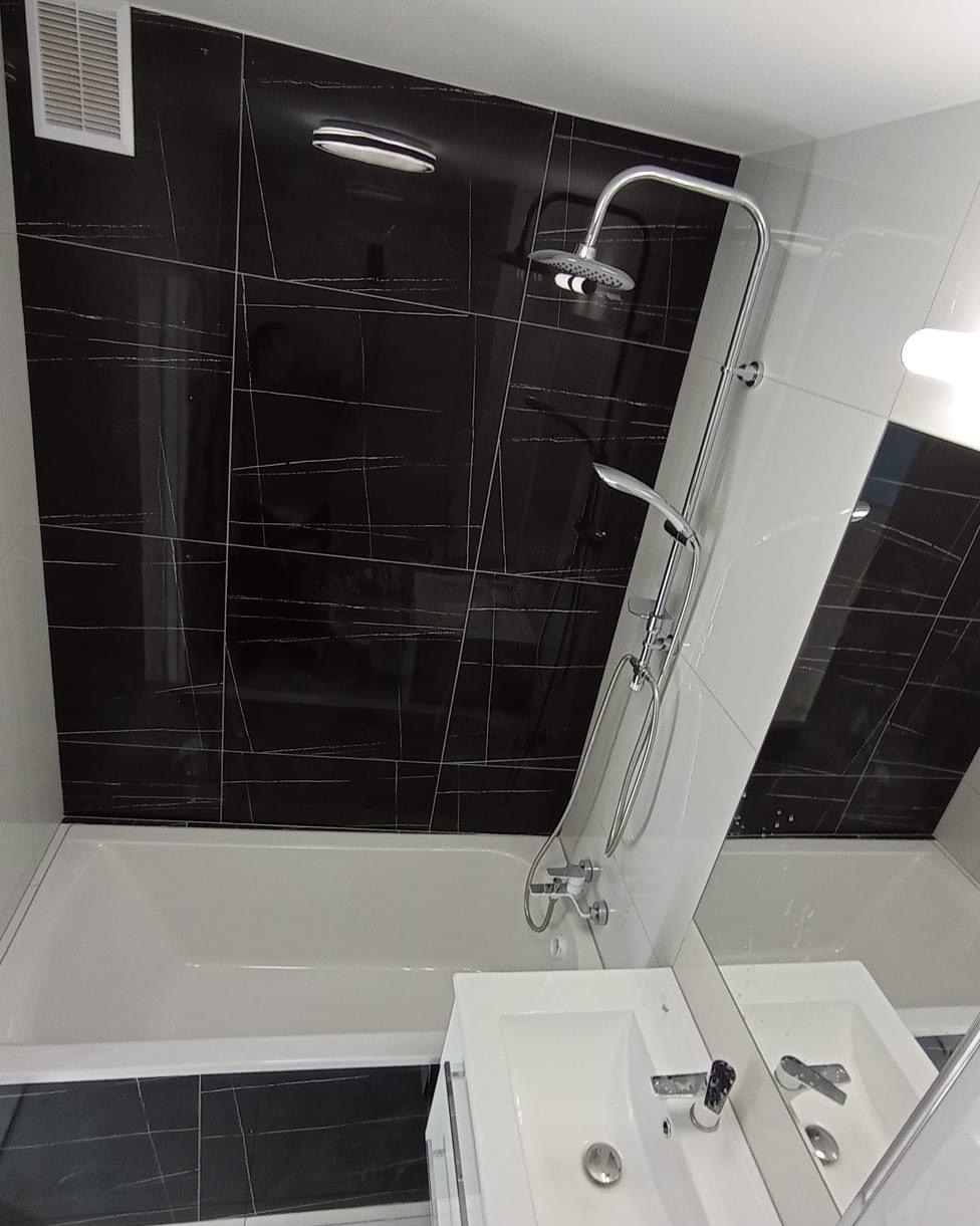 Nowoczesna łazienka z czarno-białymi płytkami, bateria prysznicowa z deszczownicą, biała wanna i umywalka z szafką, widoczny fragment wentylacji.