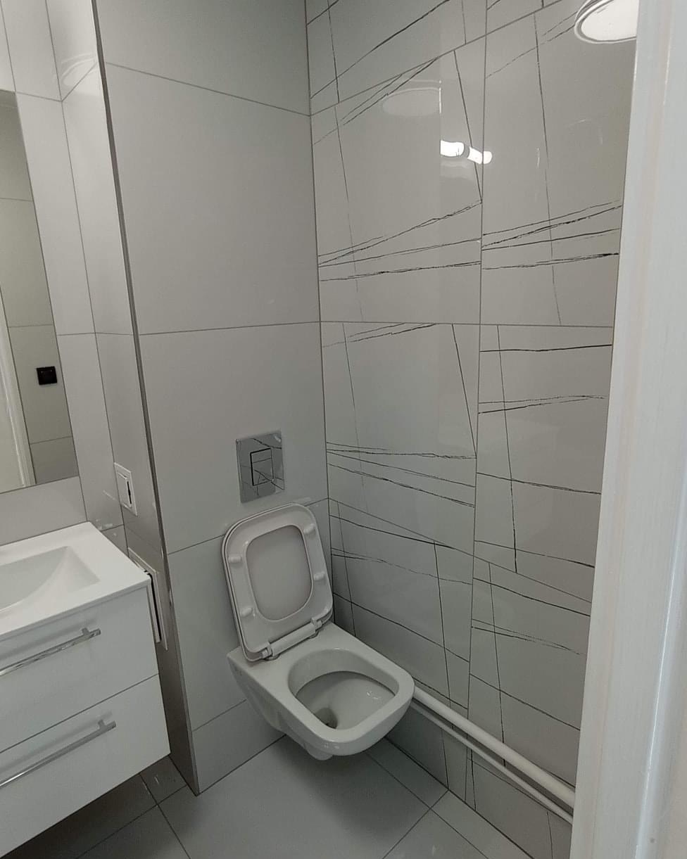 Biała wisząca toaleta z otwartą klapą w nowoczesnej łazience z białymi płytkami ozdobionymi szarymi liniami, obok umywalka z szafką i chromowanymi uchwytami.