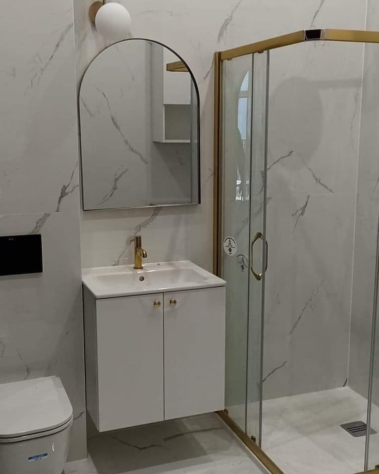 Nowoczesna łazienka z białymi marmurkowymi płytkami, kabiną prysznicową ze złotymi ramami, umywalką nablatową z szafką i lustrem w kształcie łuku.