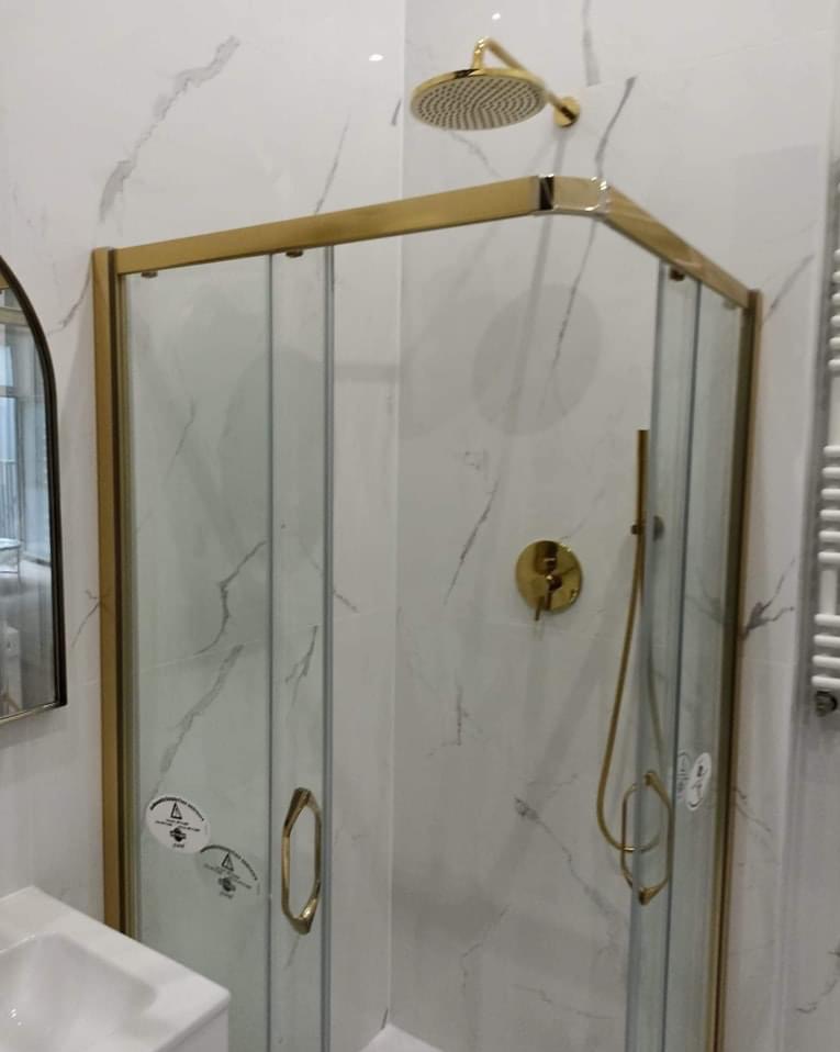 Kabina prysznicowa z złotymi elementami na tle białych marmurowych płytek, widoczna bateria i deszczownica w tym samym kolorze.
