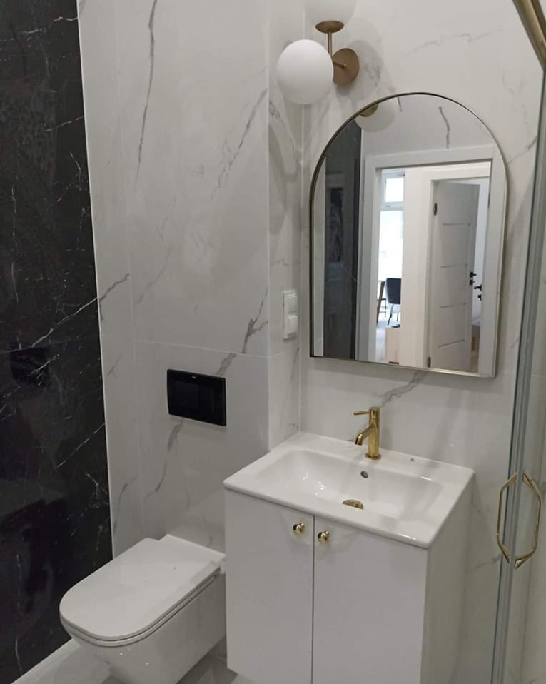 Nowoczesna łazienka z białymi marmurkowymi płytkami, czarnym akcentem, wiszącą toaletą, białą szafką z umywalką i złotą armaturą oraz lustrem w kształcie łuku.