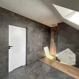 Wykończona łazienka na poddaszu z szarymi płytkami imitującymi kamień, kabiną prysznicową ze szklaną ścianką i drewnopodobną podłogą w strefie prysznica, oświetlona światłem wpadającym przez okno...