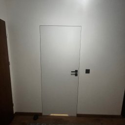 Michał Słowikowski - Minimalistyczne, białe drzwi ukryte w ścianie, z czarną klamką i włącznikiem, na tle drewnianej podłogi i wzorzystego dywanu. Nowoczesny design.