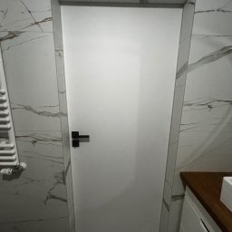 Michał Słowikowski - Białe, minimalistyczne drzwi w łazience z marmurowymi płytkami. Czarna klamka, nowoczesny grzejnik i fragment szafki umywalkowej. Wykończenie po remoncie.