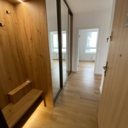 Michał Słowikowski - Nowoczesny przedpokój z drewnianą zabudową, lustrzaną szafą przesuwną i podświetlanymi półkami. Jasne panele podłogowe i widok na dalsze pomieszczenia.