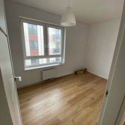 Michał Słowikowski - Jasne panele podłogowe w pokoju z oknem i grzejnikiem. Widok zza uchylonych drzwi, pudełko przy ścianie. Nowe budownictwo, minimalistyczny styl.