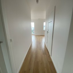 Michał Słowikowski - Jasne panele podłogowe w korytarzu, świeżo po remoncie w Szczecinie. Białe ściany i drzwi, widok na okno w oddali. Minimalistyczny design.