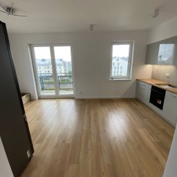 Michał Słowikowski - Jasne panele podłogowe w nowym mieszkaniu z balkonem i kuchnią w odcieniach szarości. Widok na osiedle przez okno balkonowe. Minimalistyczny design.