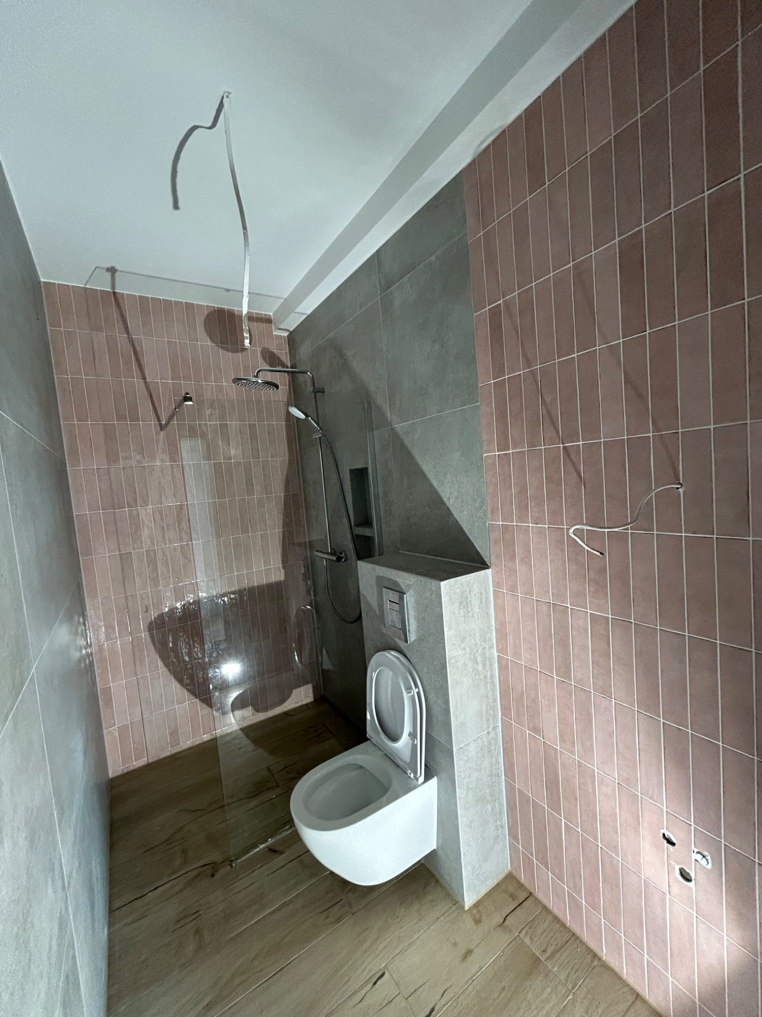 Wnętrze łazienki w trakcie wykańczania, widoczne płytki w odcieniach różu i szarości, drewnopodobna podłoga, biała toaleta oraz kabina prysznicowa z zamontowaną deszczownicą, wystające kable...