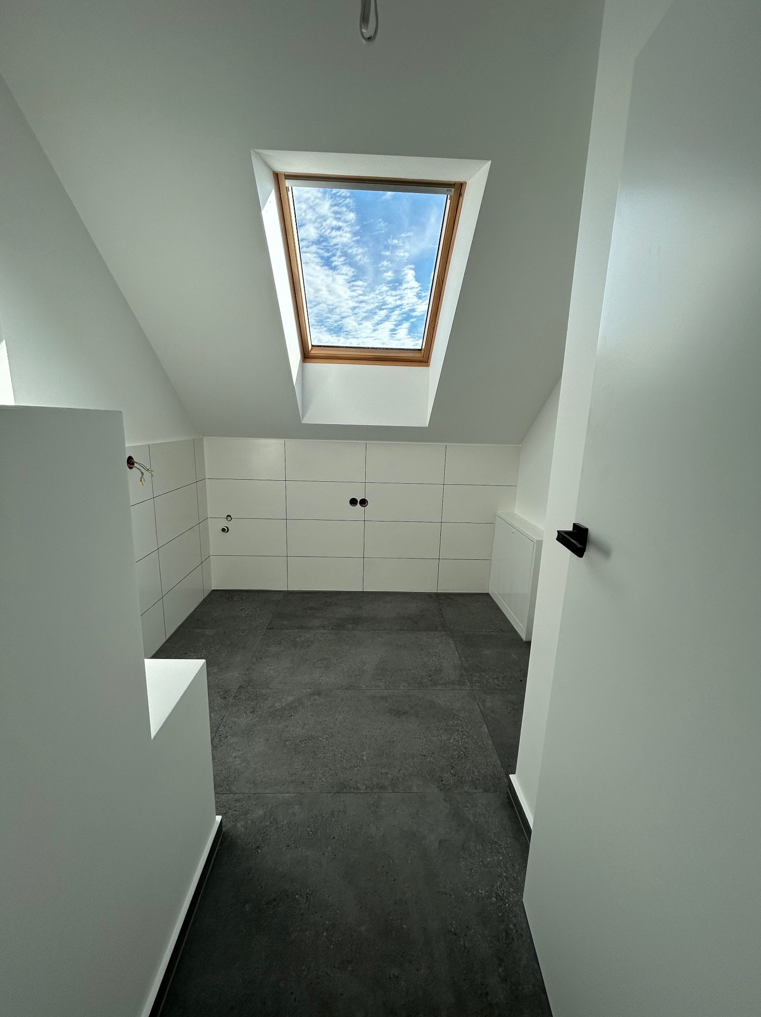 Wnętrze poddasza ze świeżo ułożonymi, kwadratowymi, białymi płytkami na ścianie, ciemnoszarą podłogą i oknem dachowym, przez które widać błękitne niebo z chmurami. Widoczne puszki elektryczne...