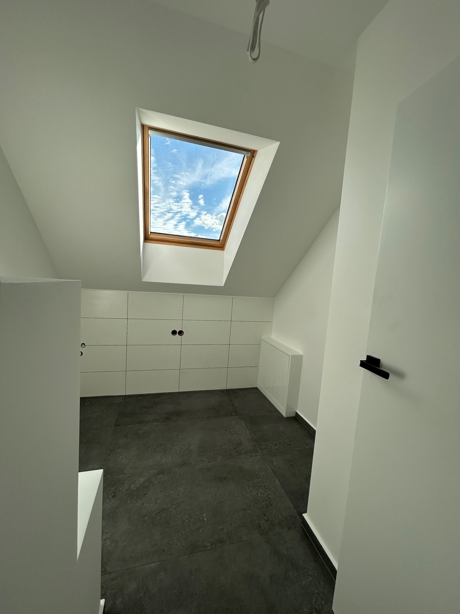 Wnętrze poddasza ze skośnym sufitem, wykończone białymi ścianami i szarą podłogą z płytek. Widoczne okno dachowe z drewnianą ramą, przez które widać błękitne niebo z chmurami. Na ścianie białe...