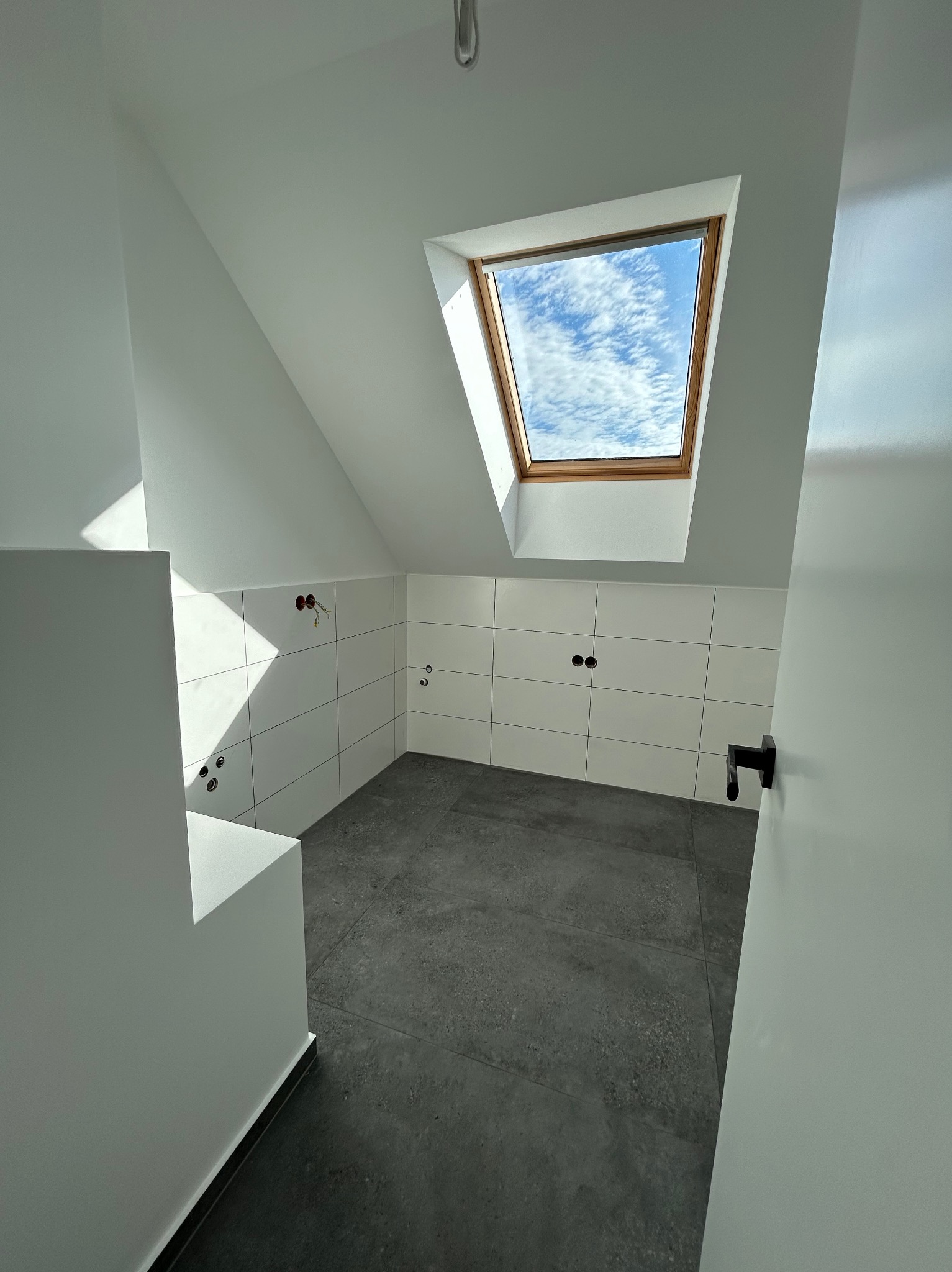 Surowe wnętrze łazienki na poddaszu z oknem dachowym, wyłożone białymi płytkami i szarą podłogą, widoczne wystające kable elektryczne i brakujące elementy armatury.