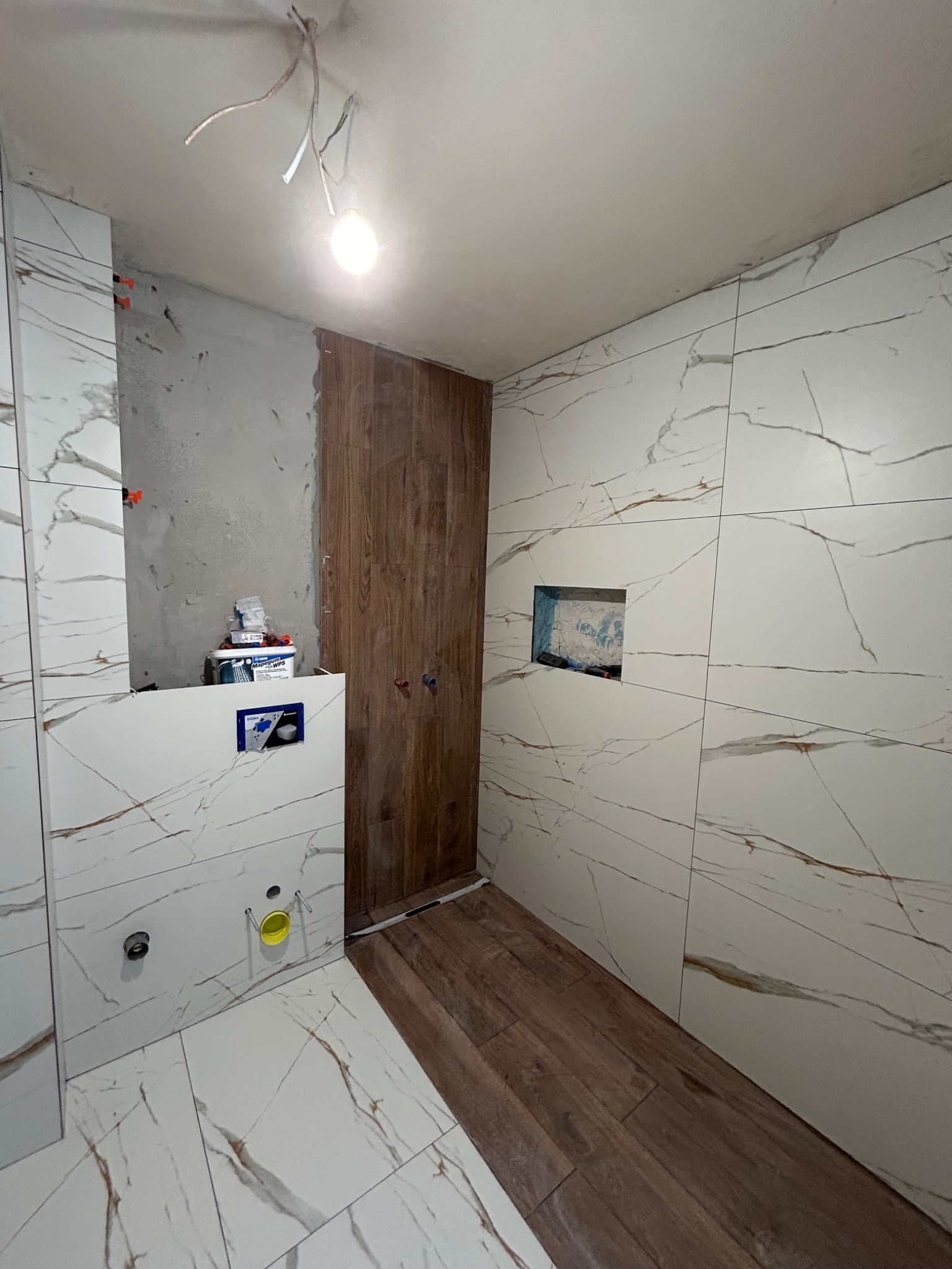 Wnętrze łazienki w trakcie wykańczania: częściowo ułożone płytki imitujące marmur na ścianach i podłodze, fragment ściany z drewnopodobnymi panelami, widoczne puszki instalacyjne i pojemnik...