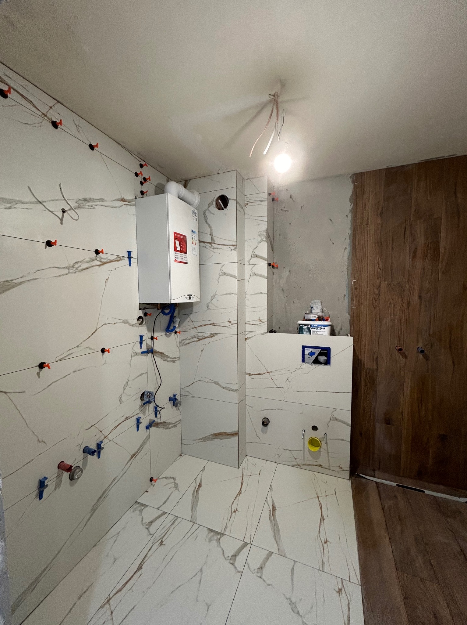 Wnętrze łazienki w trakcie remontu, z białymi płytkami imitującymi marmur na ścianach i podłodze, system poziomowania płytek z klinami i krzyżykami, zamontowany piec gazowy, częściowo wykończona...