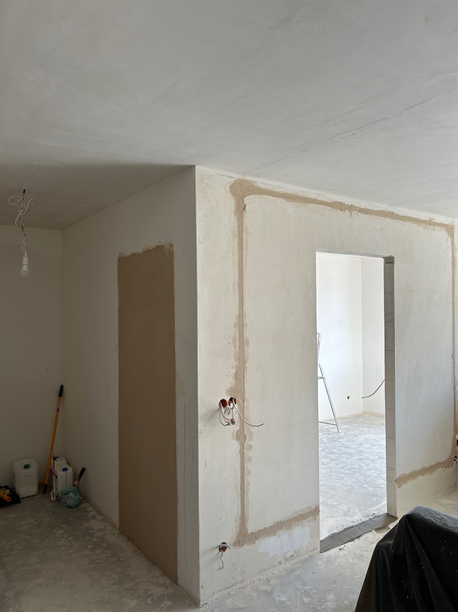 Wnętrze w trakcie prac wykończeniowych: ściany po nałożeniu gładzi, widoczne przewody elektryczne, częściowo zakryta podłoga, drabina w tle, przygotowane narzędzia i materiały budowlane w rogu...