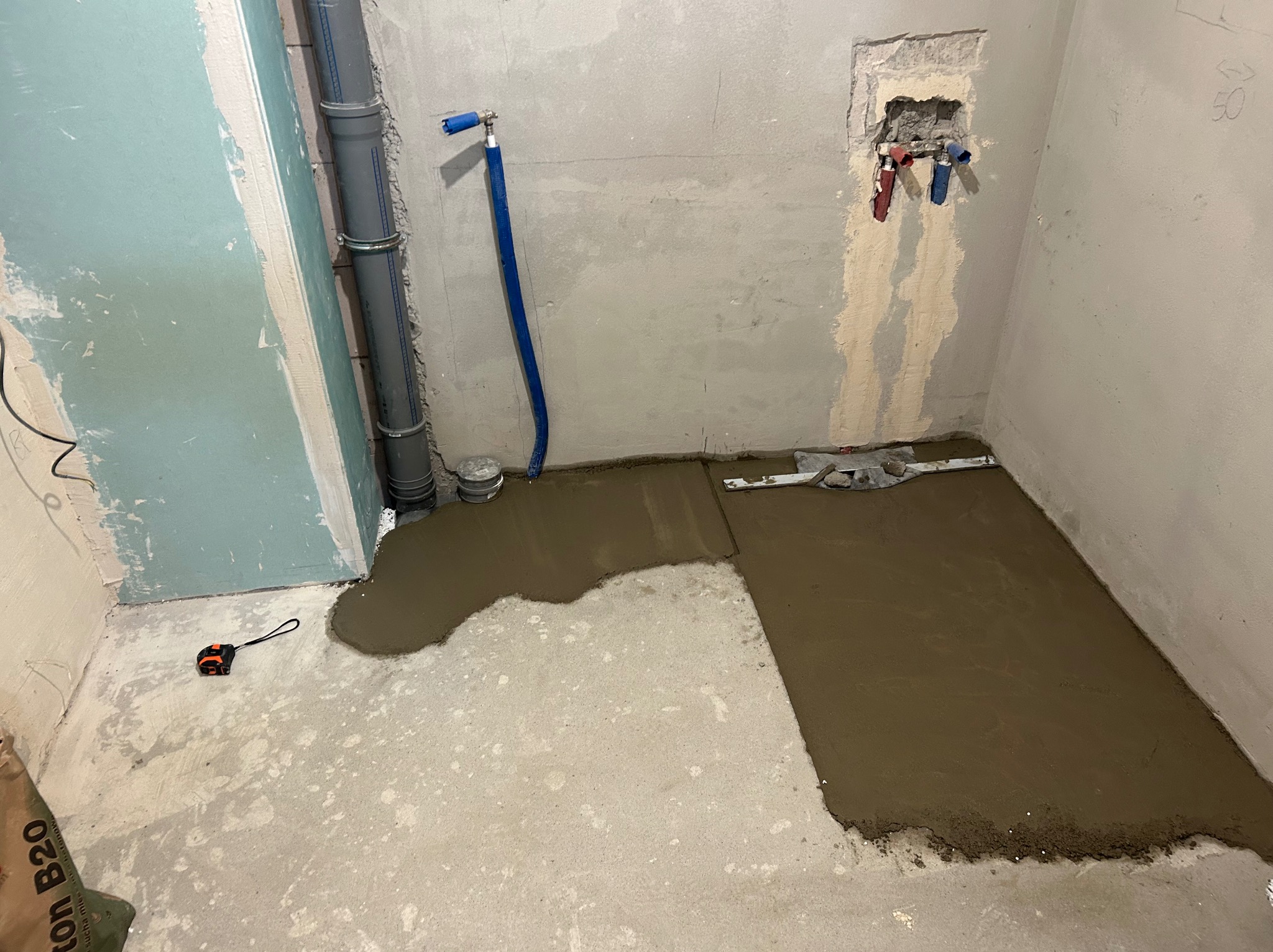 Wyrównywanie podłoża w łazience za pomocą wylewki samopoziomującej; widoczna poziomica, rury instalacyjne i worek z zaprawą.