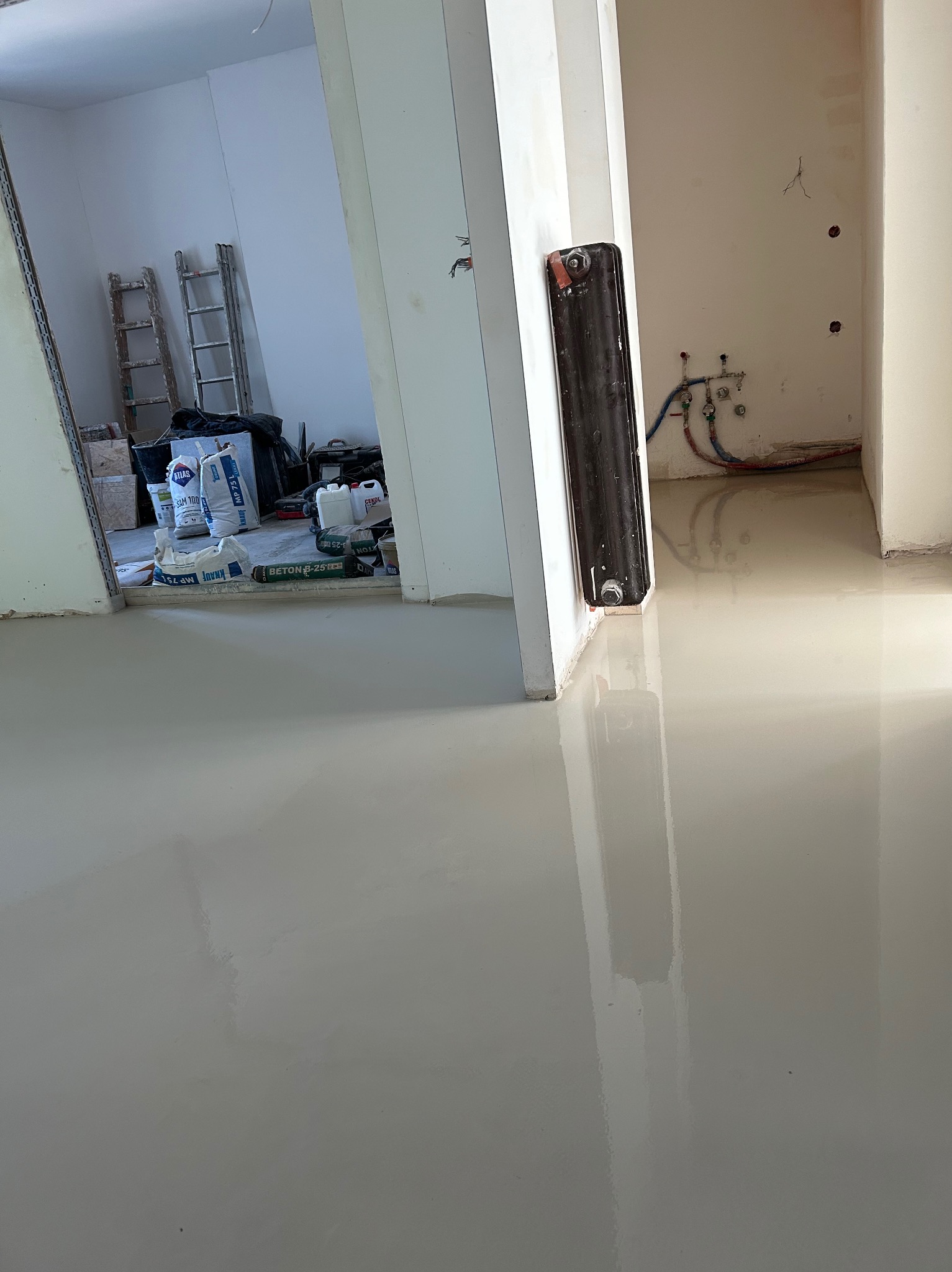 Wylewka samopoziomująca na podłodze w trakcie realizacji, widoczne worki z materiałami budowlanymi Knauf MP 75, Atlas SAM 100 i Beton B-25 oraz drabiny w tle.