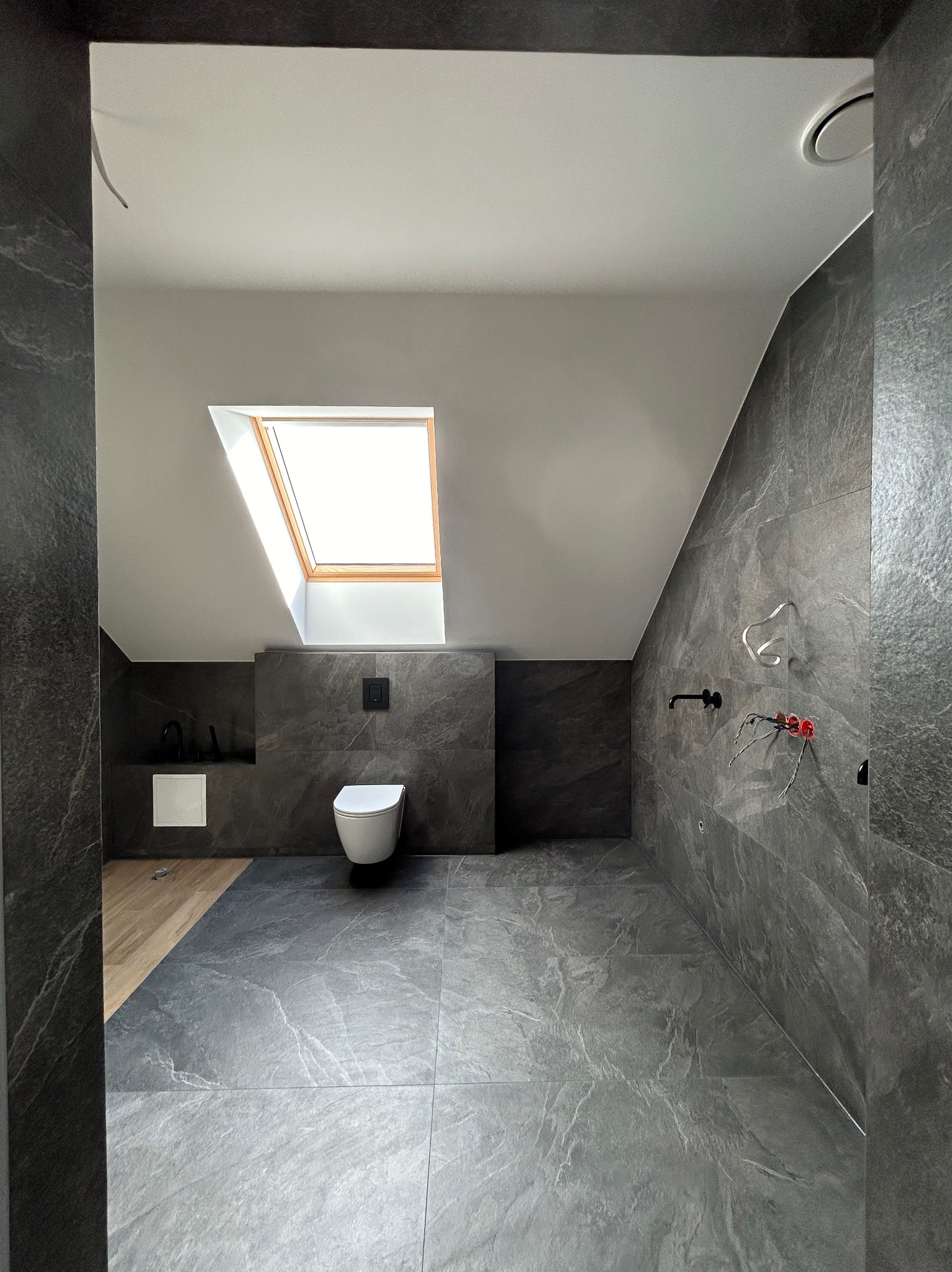 Wykończona łazienka na poddaszu w szarościach z drewnianym akcentem pod umywalką, widoczne przyłącza wodne na ścianie, oświetlona przez okno dachowe.