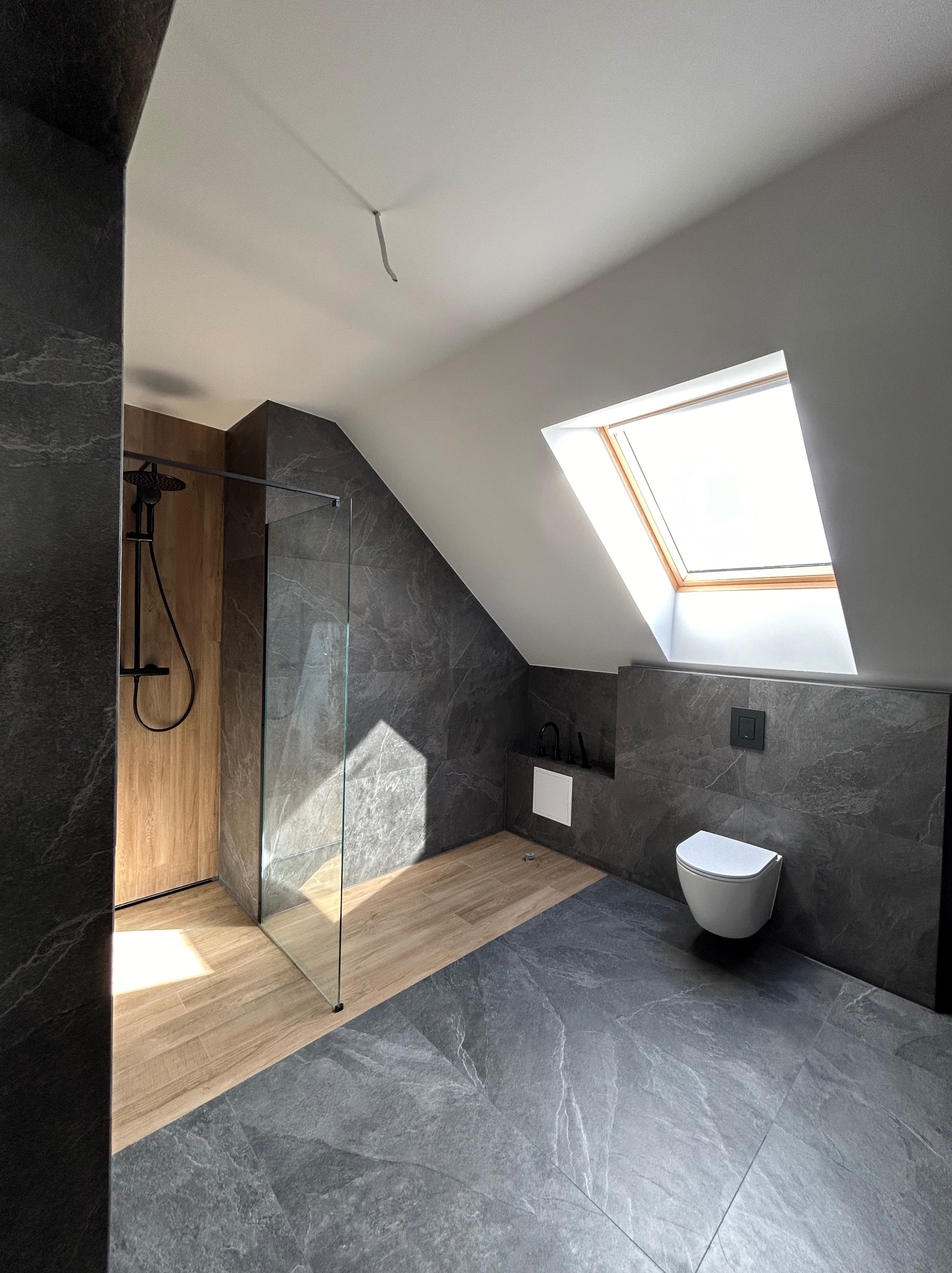 Nowoczesna łazienka na poddaszu z kabiną prysznicową typu walk-in, wykończona szarymi płytkami imitującymi kamień i drewnopodobnymi panelami podłogowymi, oświetlona światłem dziennym wpadającym...