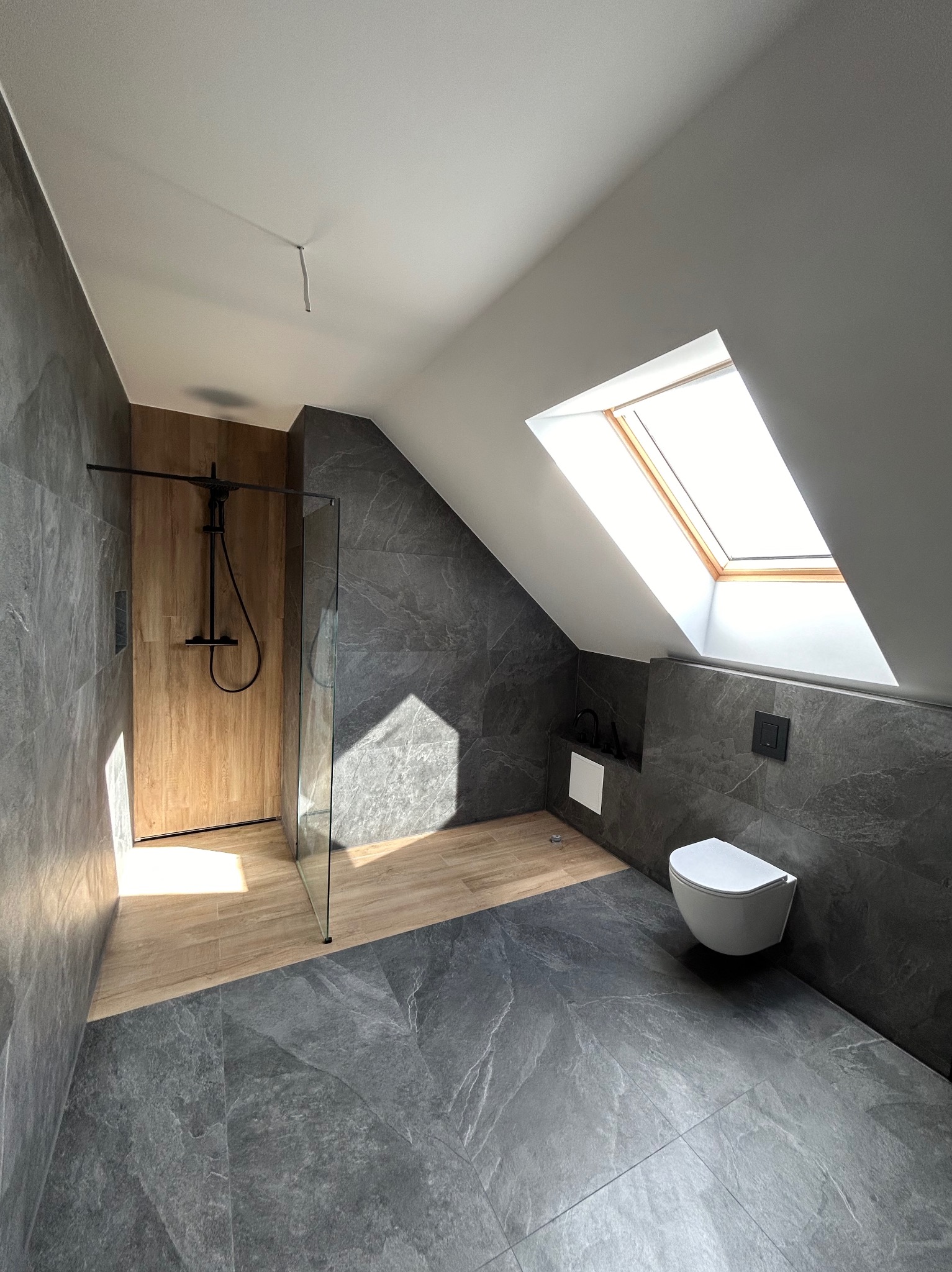 Nowoczesna łazienka na poddaszu z szarymi płytkami imitującymi kamień, kabiną prysznicową typu walk-in z drewnopodobnymi panelami na ścianie, toaletą podwieszaną i oknem dachowym wpuszczającym...