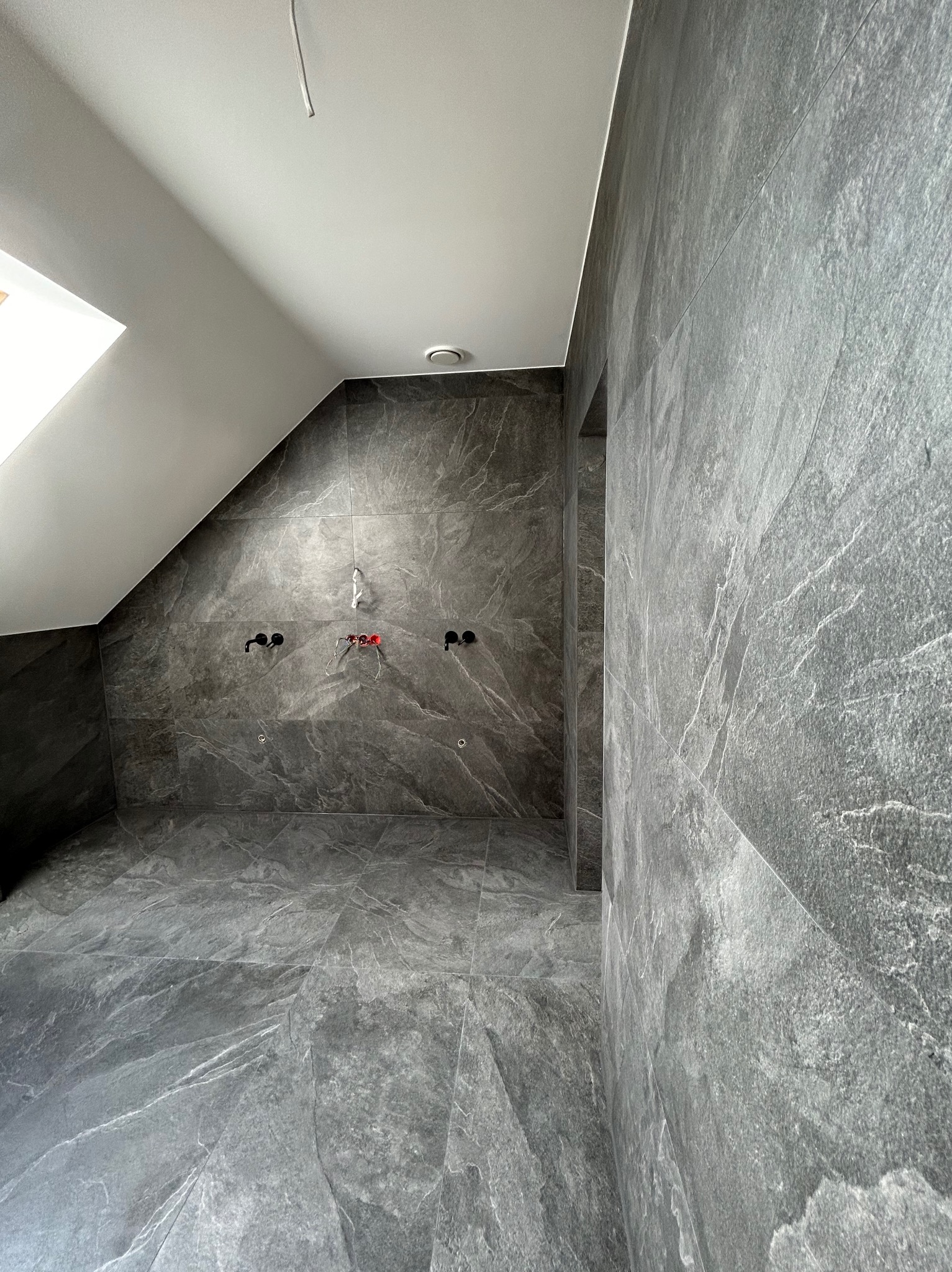 Wnętrze łazienki na poddaszu w trakcie prac wykończeniowych, widoczne szare płytki imitujące kamień na ścianach i podłodze, przygotowane przyłącza wodno-kanalizacyjne oraz surowe wykończenie sufitu.