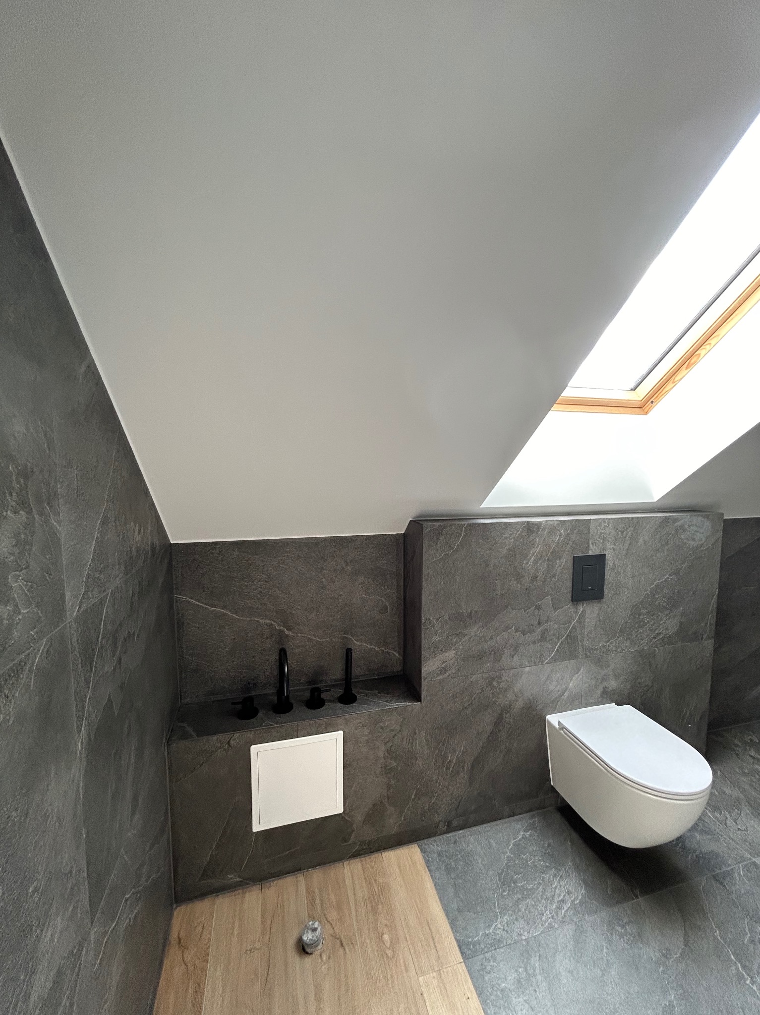 Nowoczesna łazienka na poddaszu z szarą okładziną imitującą kamień, wiszącą toaletą i drewnianym akcentem pod szafką z czarną armaturą, doświetlona oknem dachowym.