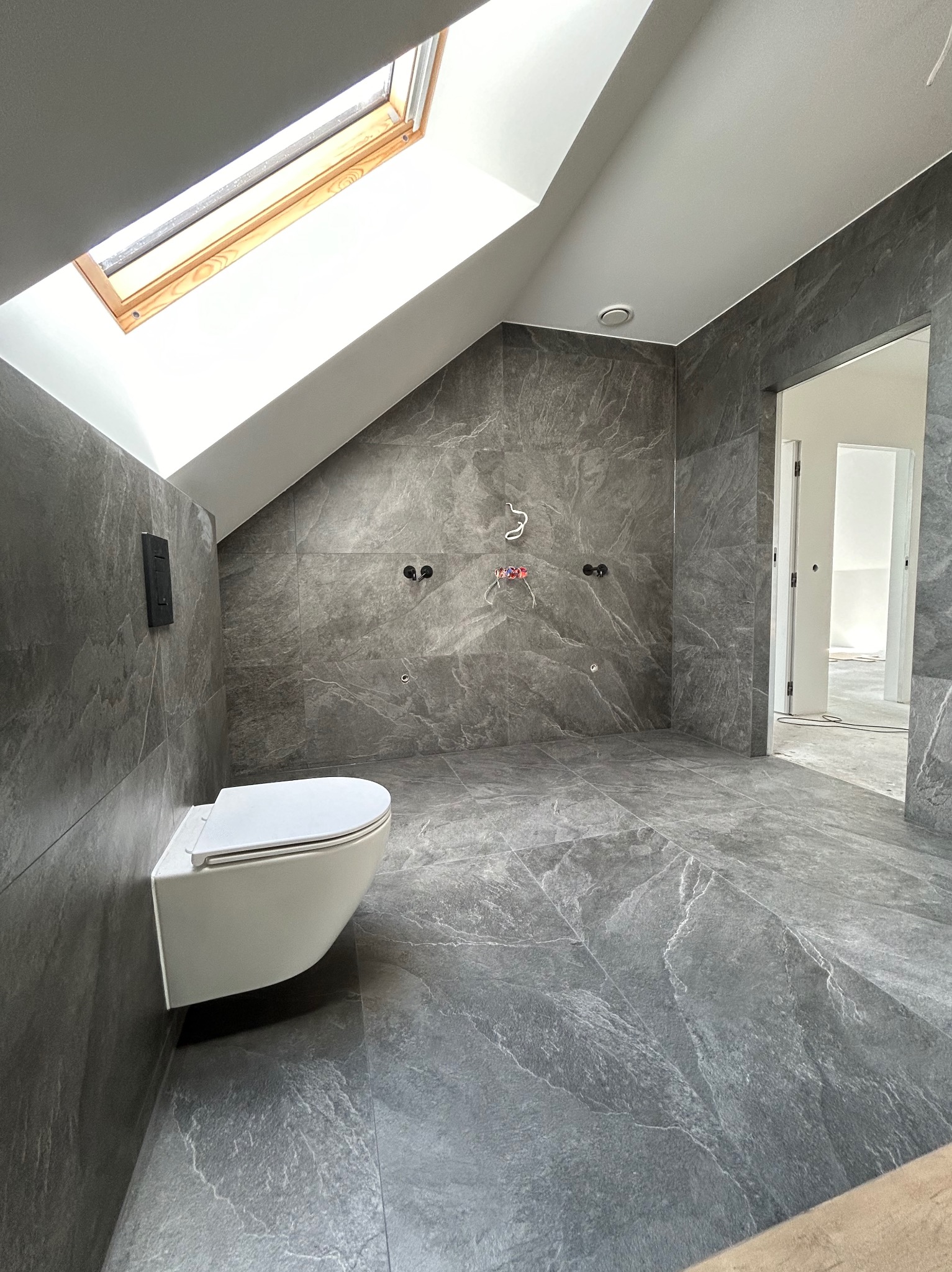 Nowoczesna łazienka na poddaszu z szarymi płytkami imitującymi kamień, wisząca toaleta, okno dachowe i instalacja wodno-kanalizacyjna w ścianie przed montażem armatury.