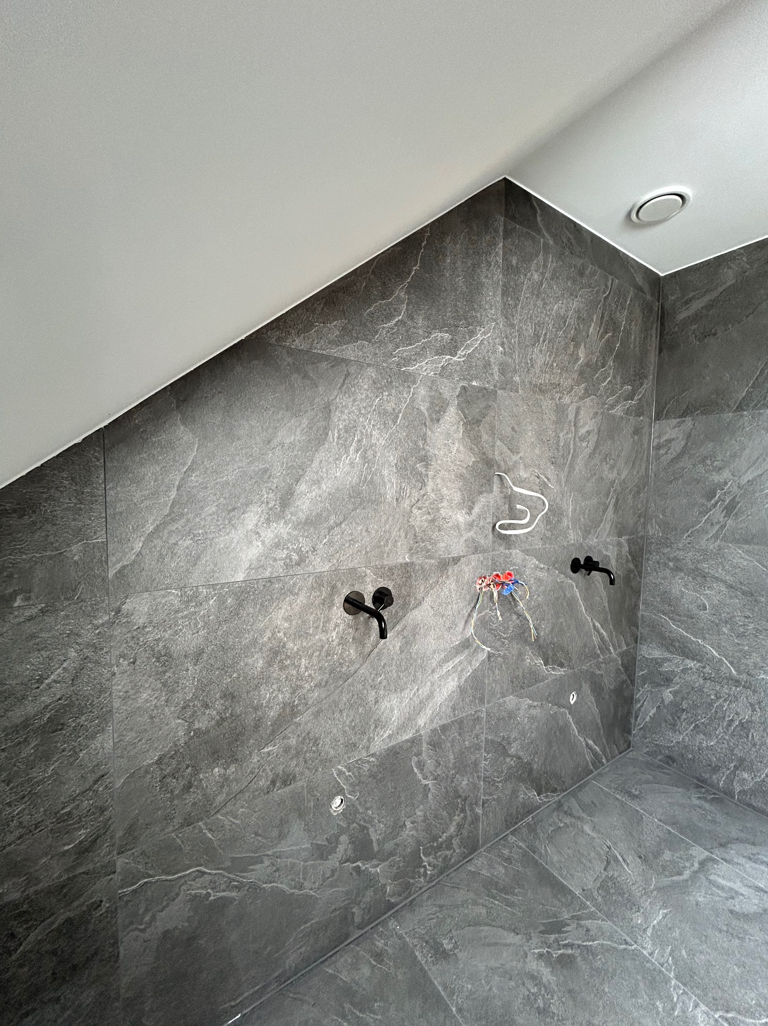 Szczecin: Ściana i podłoga łazienki wyłożone szarymi płytkami imitującymi kamień, z widocznymi czarnymi bateriami, przewodami elektrycznymi i wieszakiem na ręcznik, w trakcie montażu.