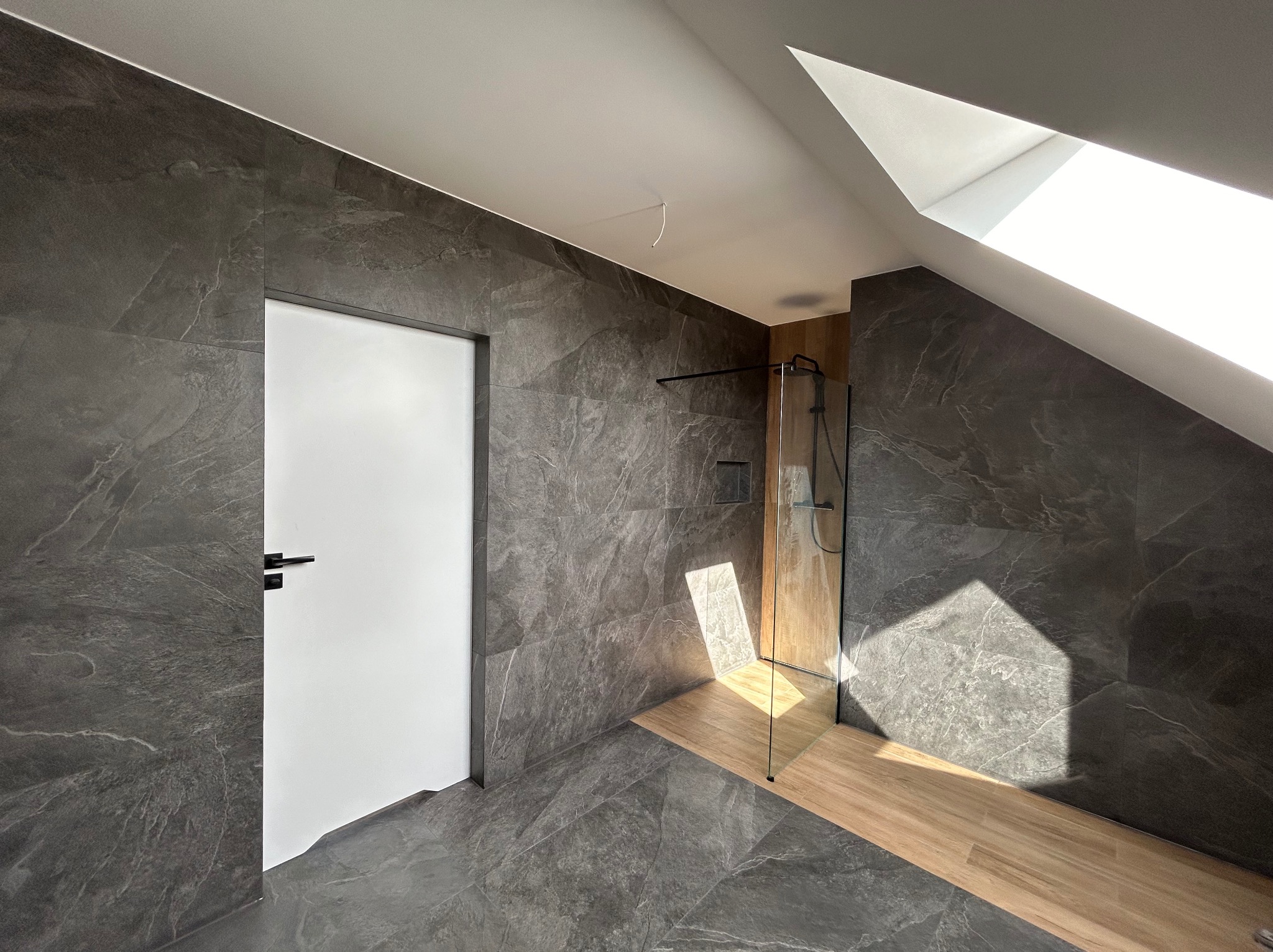 Wykończona łazienka na poddaszu z szarymi płytkami imitującymi kamień, kabiną prysznicową ze szklaną ścianką i drewnopodobną podłogą w strefie prysznica, oświetlona światłem wpadającym przez okno...