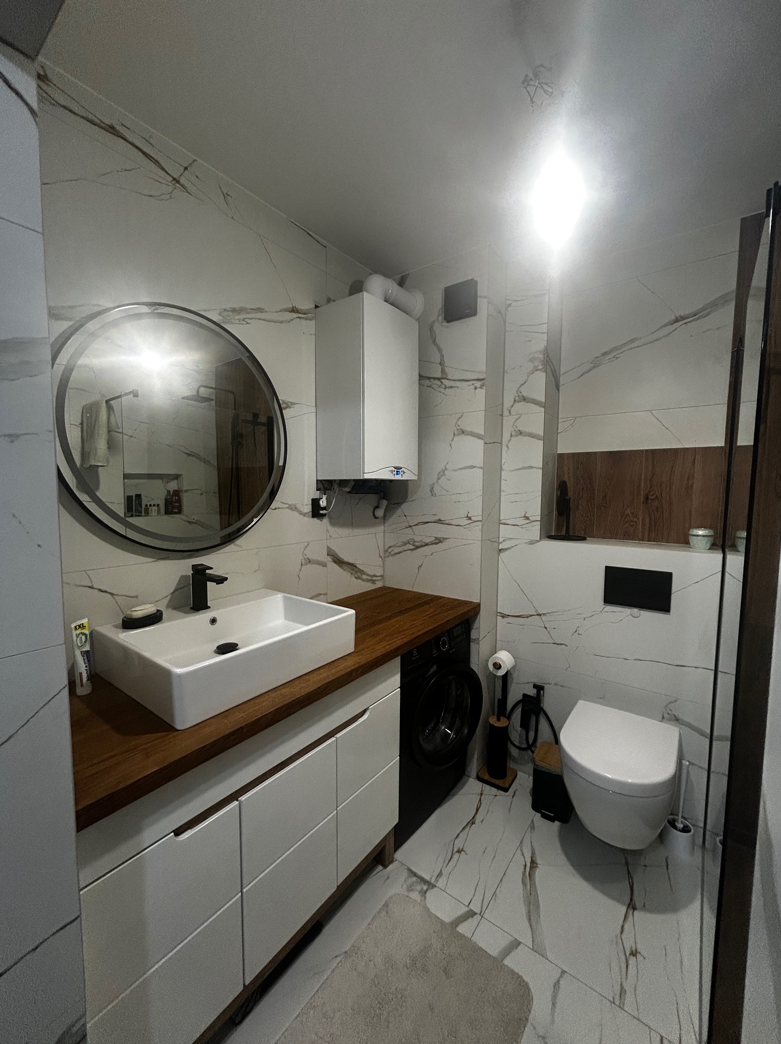 Nowoczesna łazienka z umywalką nablatową, okrągłym lustrem i płytkami imitującymi marmur. Widoczny bojler, pralka i toaleta. Całość utrzymana w jasnej kolorystyce.