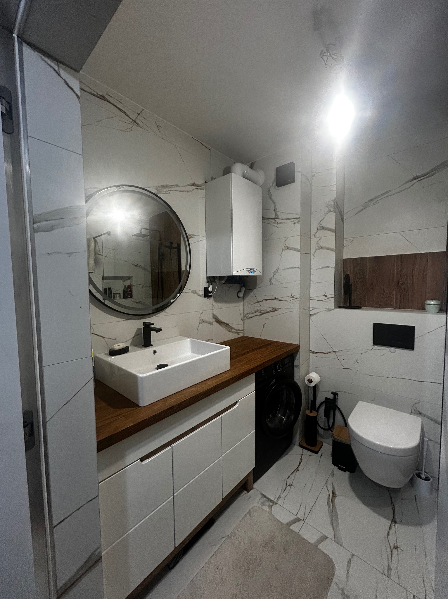 Nowoczesna łazienka z marmurowymi płytkami, drewnianym blatem pod umywalką i okrągłym lustrem. Widoczna pralka i wisząca toaleta.