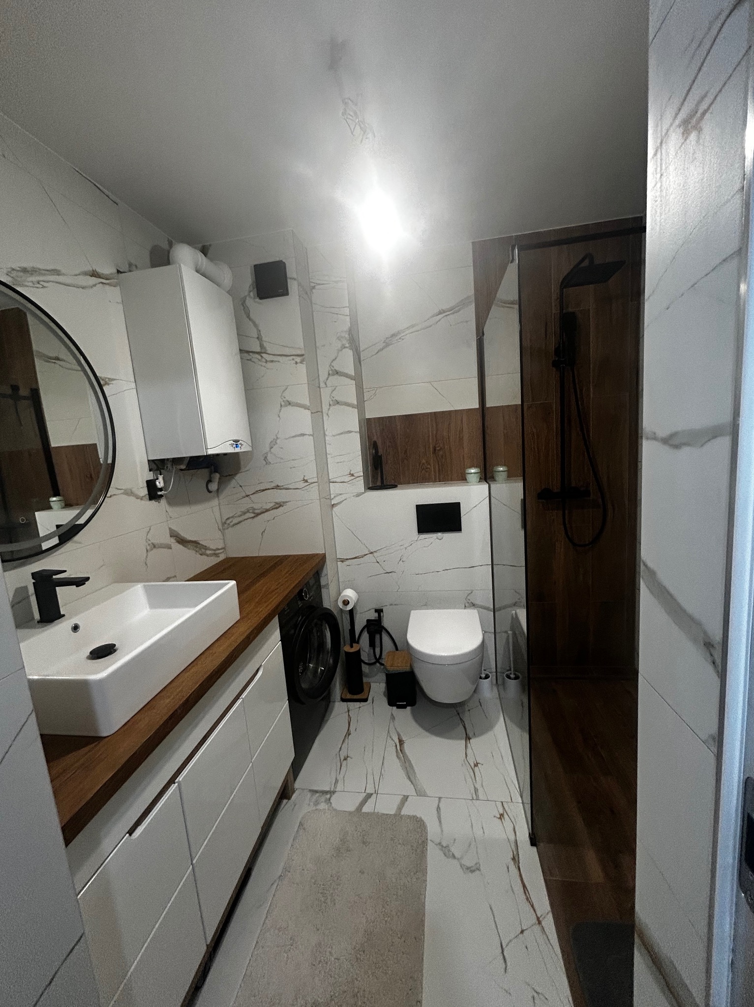 Nowoczesna łazienka z marmurowymi płytkami, drewnianym blatem i czarną armaturą. Kabina prysznicowa z ciemnego drewna, okrągłe lustro i biała umywalka.