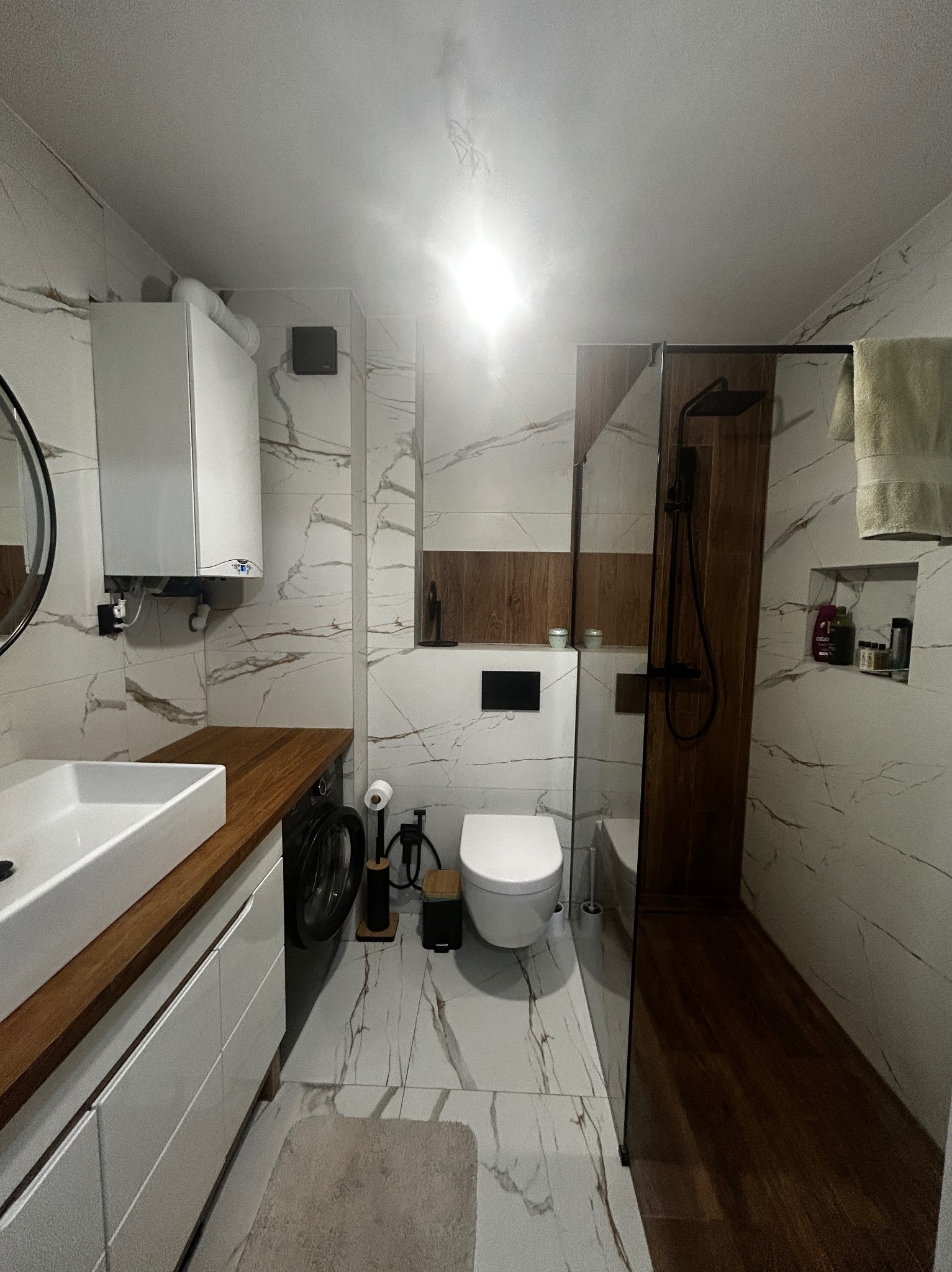 Nowoczesna łazienka po remoncie w Szczecinie: białe marmurowe płytki, drewniane akcenty, kabina prysznicowa typu walk-in i umywalka z blatem z drewna.