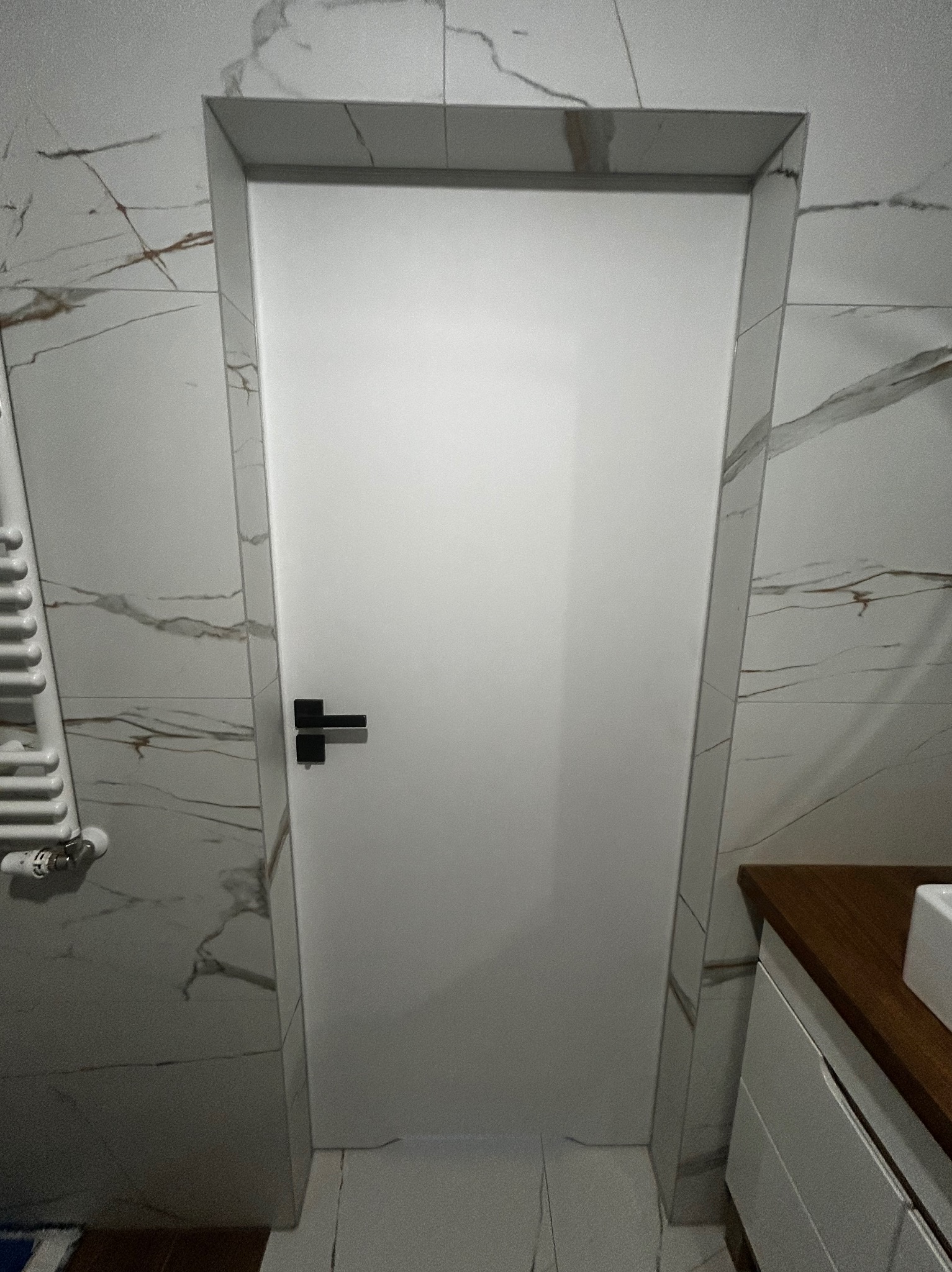 Białe, minimalistyczne drzwi w łazience z marmurowymi płytkami. Czarna klamka, nowoczesny grzejnik i fragment szafki umywalkowej. Wykończenie po remoncie.