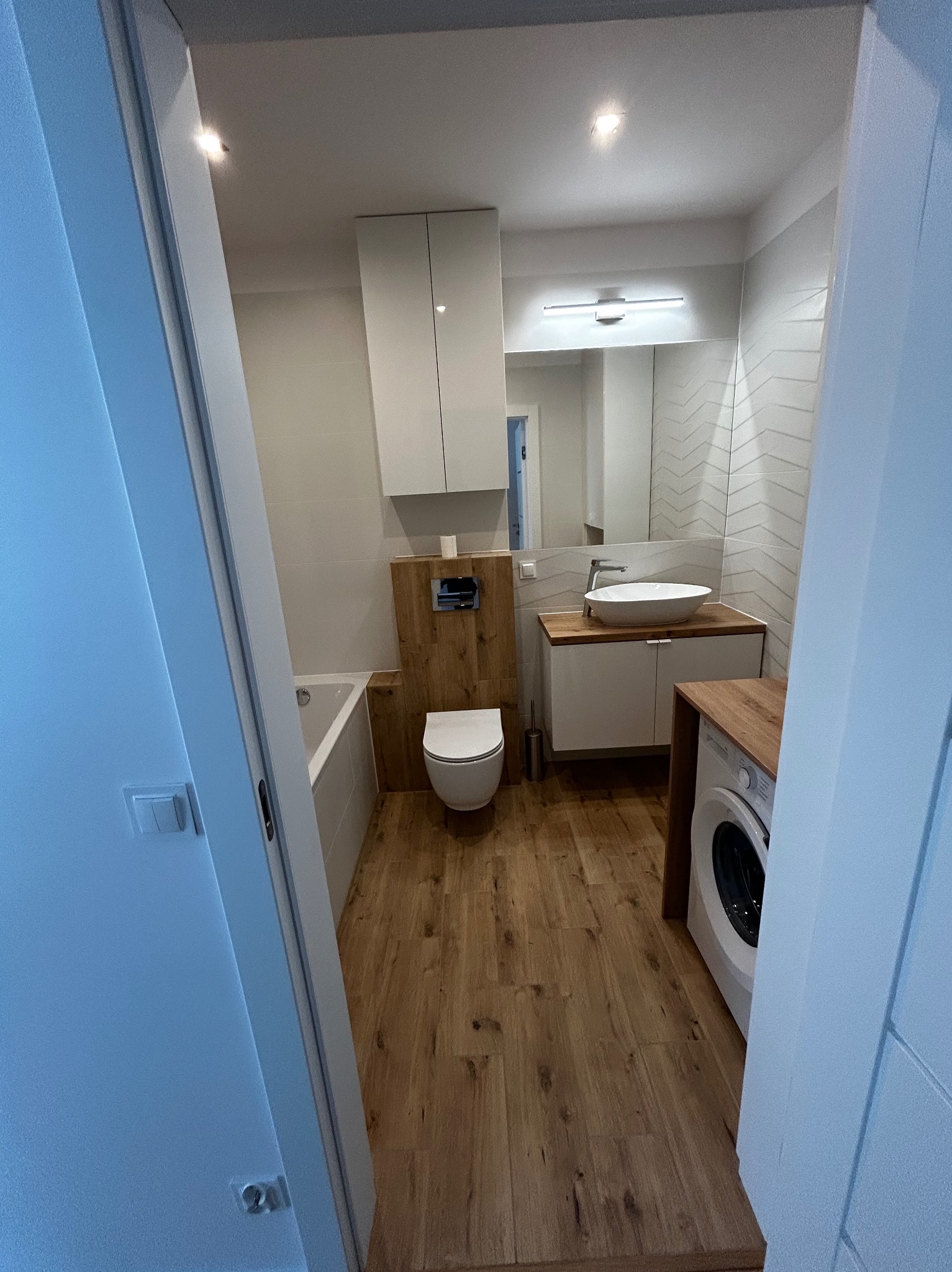 Nowoczesna łazienka z drewnianą podłogą i akcentami, białą armaturą i pralką. Umywalka nablatowa, lustro z oświetleniem. Widok zza otwartych drzwi.