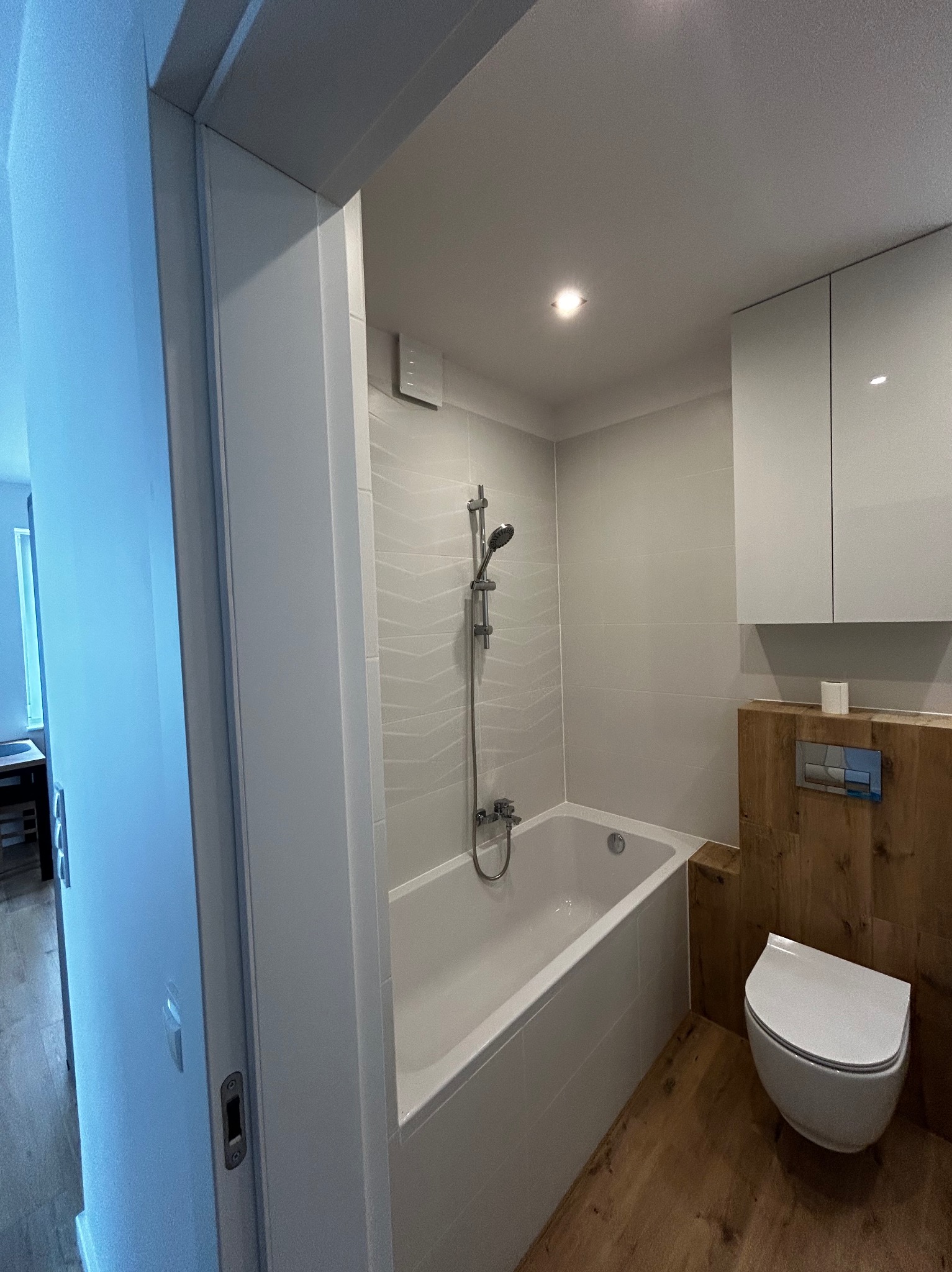 Nowoczesna łazienka po remoncie: biała wanna z prysznicem, podwieszana toaleta, drewniane akcenty i subtelne oświetlenie. Widok zza otwartych drzwi.