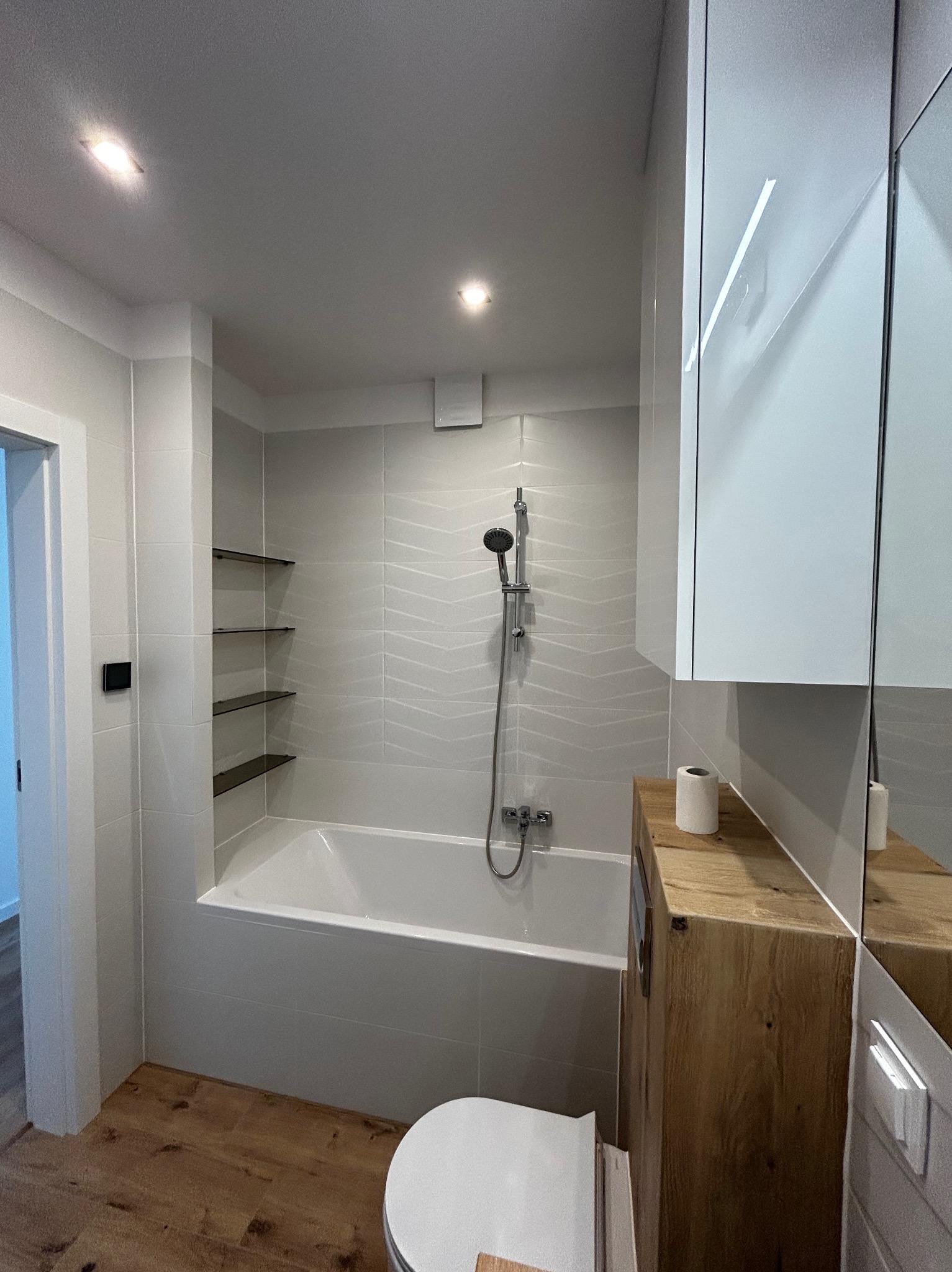 Nowoczesna łazienka po remoncie: białe płytki z subtelnym wzorem, drewnopodobna zabudowa toalety, wanna z prysznicem i szklane półki wnękowe.