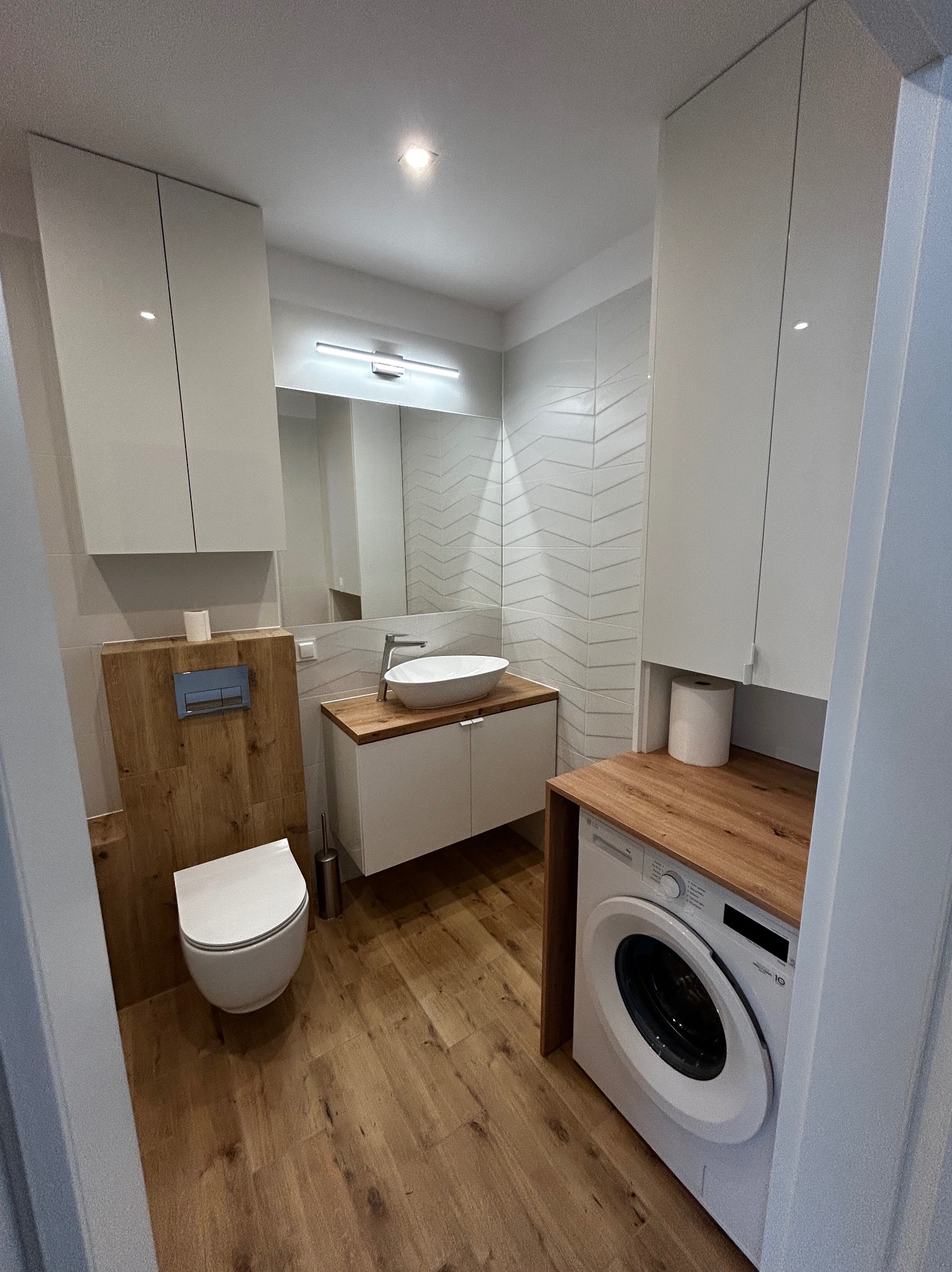 Nowoczesna łazienka po renowacji: drewniana podłoga, białe meble, umywalka nablatowa, pralka pod blatem. Ściany z białymi płytkami w geometryczny wzór.