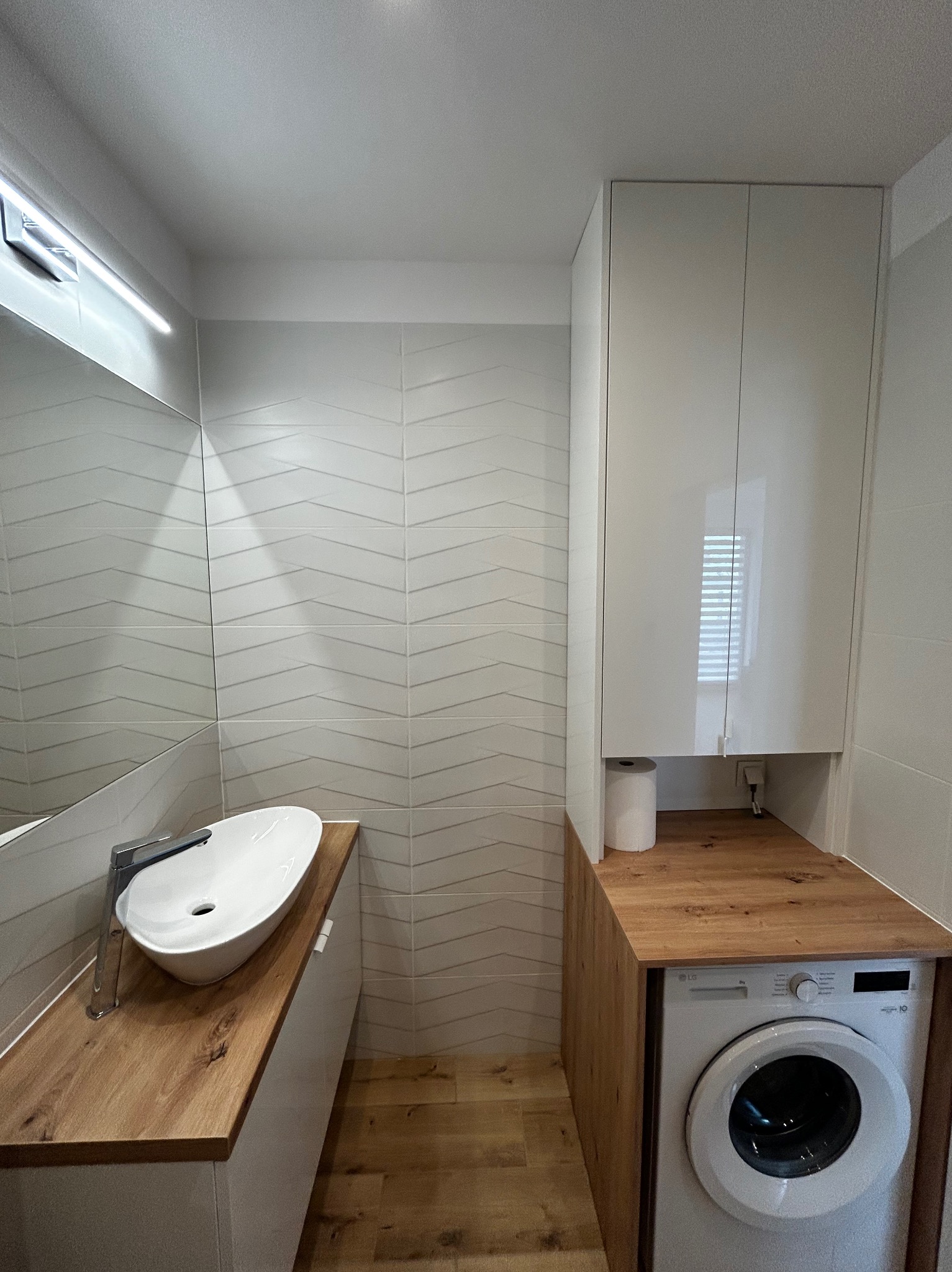 Nowoczesna łazienka po remoncie: białe płytki z wzorem chevron, umywalka nablatowa na drewnianym blacie, pralka zabudowana pod blatem. Minimalistyczny design.