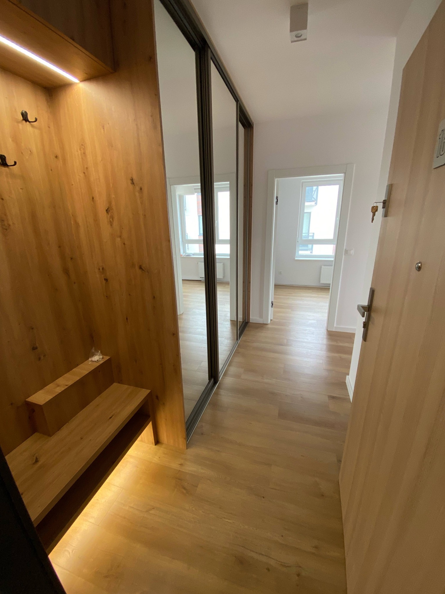 Nowoczesny przedpokój z drewnianą zabudową, lustrzaną szafą przesuwną i podświetlanymi półkami. Jasne panele podłogowe i widok na dalsze pomieszczenia.