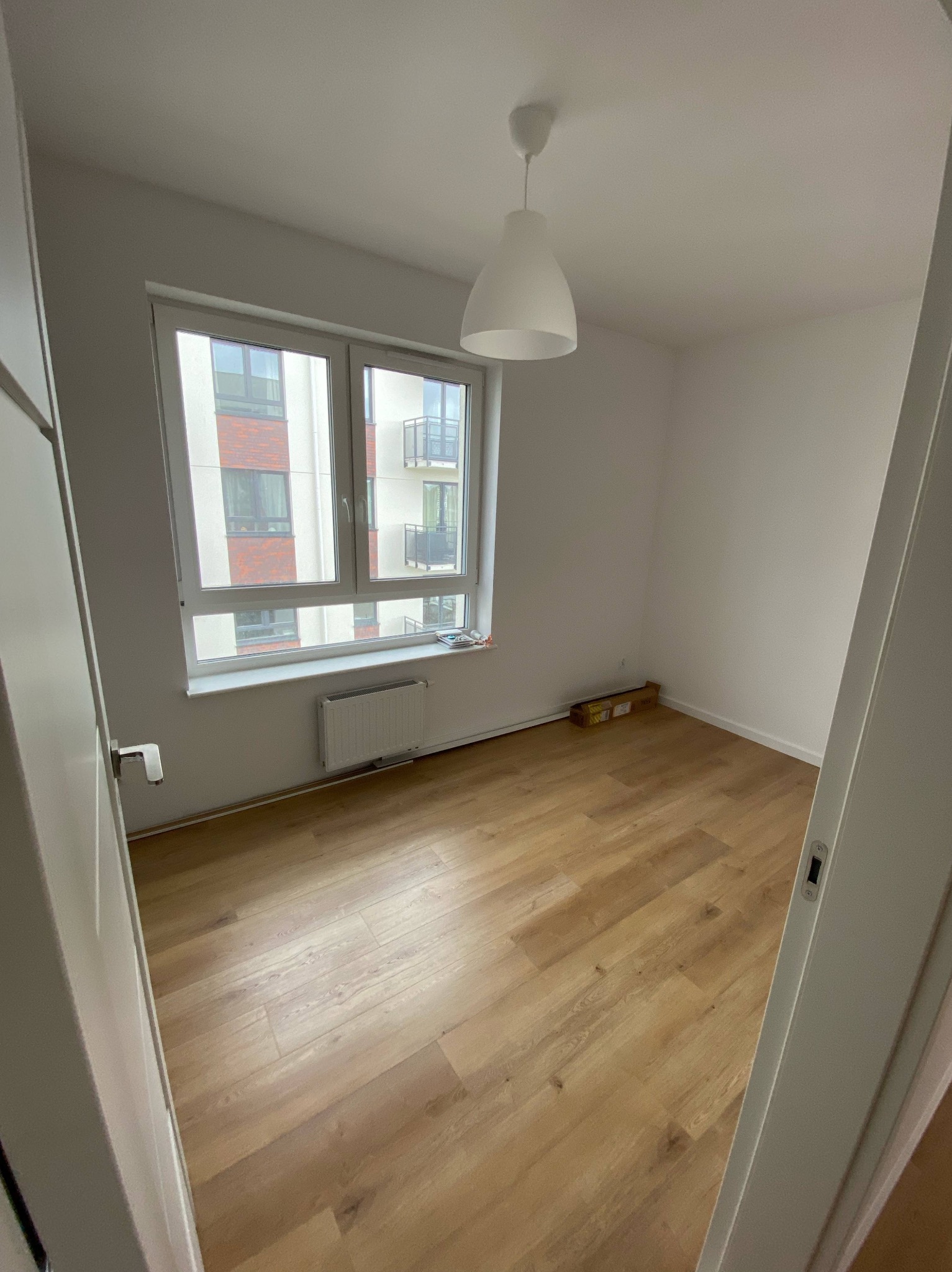 Jasne panele podłogowe w pokoju z oknem i grzejnikiem. Widok zza uchylonych drzwi, pudełko przy ścianie. Nowe budownictwo, minimalistyczny styl.