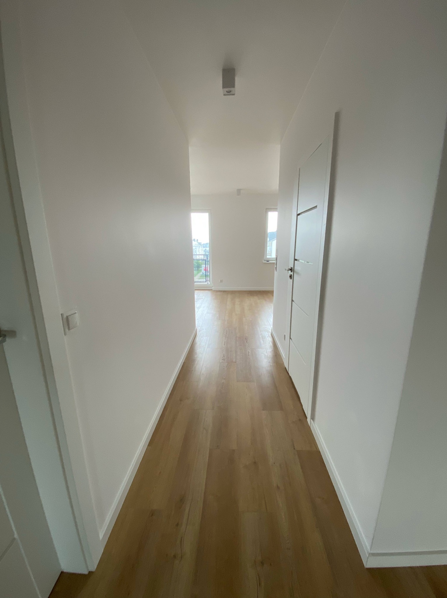 Jasne panele podłogowe w korytarzu, świeżo po remoncie w Szczecinie. Białe ściany i drzwi, widok na okno w oddali. Minimalistyczny design.