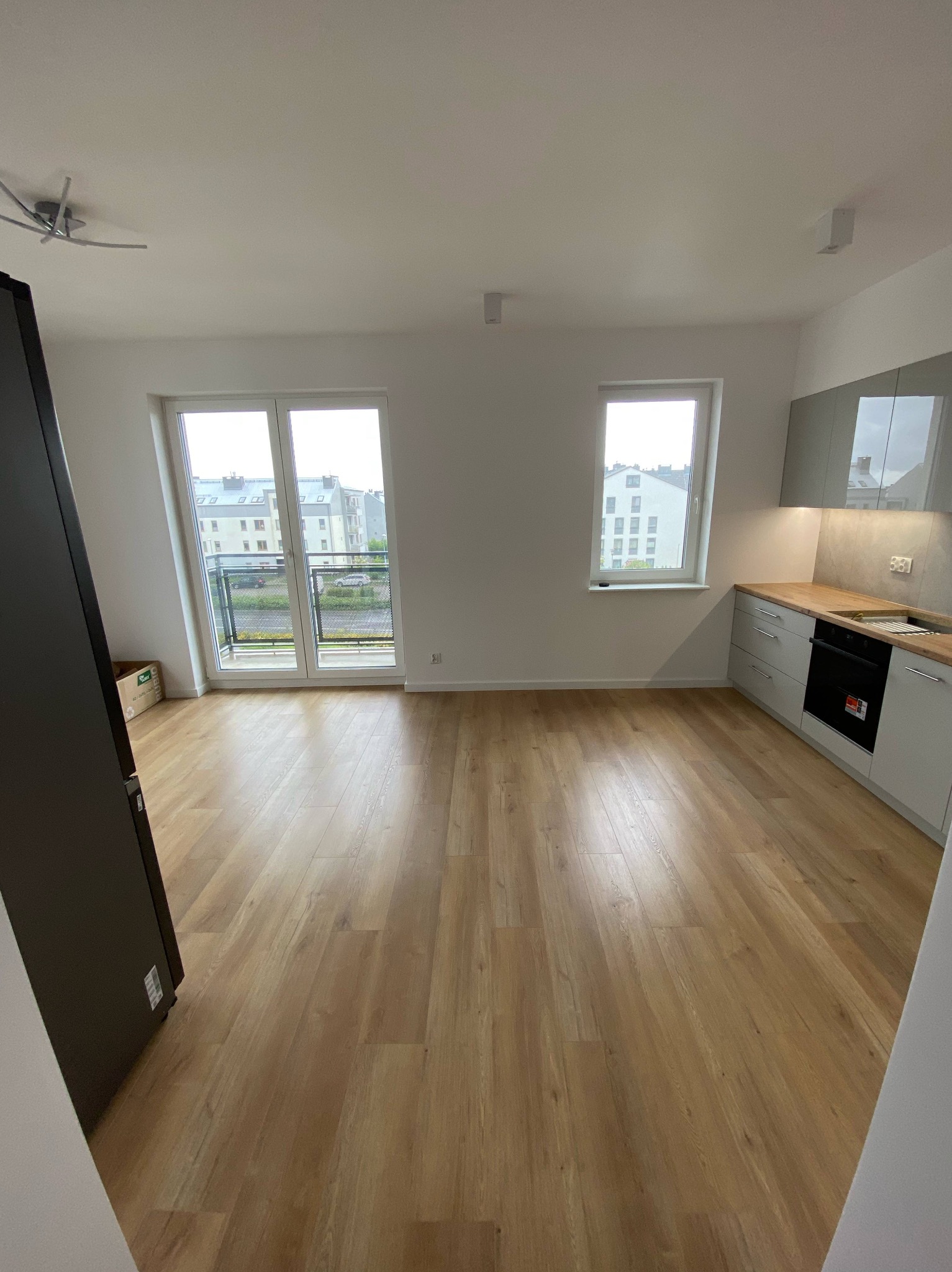 Jasne panele podłogowe w nowym mieszkaniu z balkonem i kuchnią w odcieniach szarości. Widok na osiedle przez okno balkonowe. Minimalistyczny design.