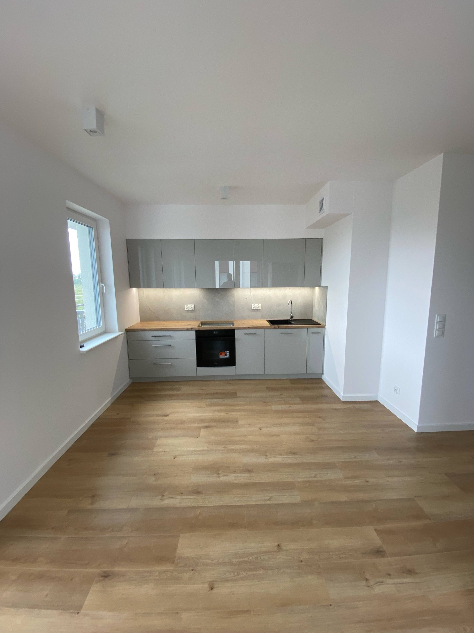 Jasna kuchnia z szarymi szafkami i drewnianym blatem, oświetlona listwą LED, z drewnianą podłogą i białymi ścianami. Minimalistyczny design.