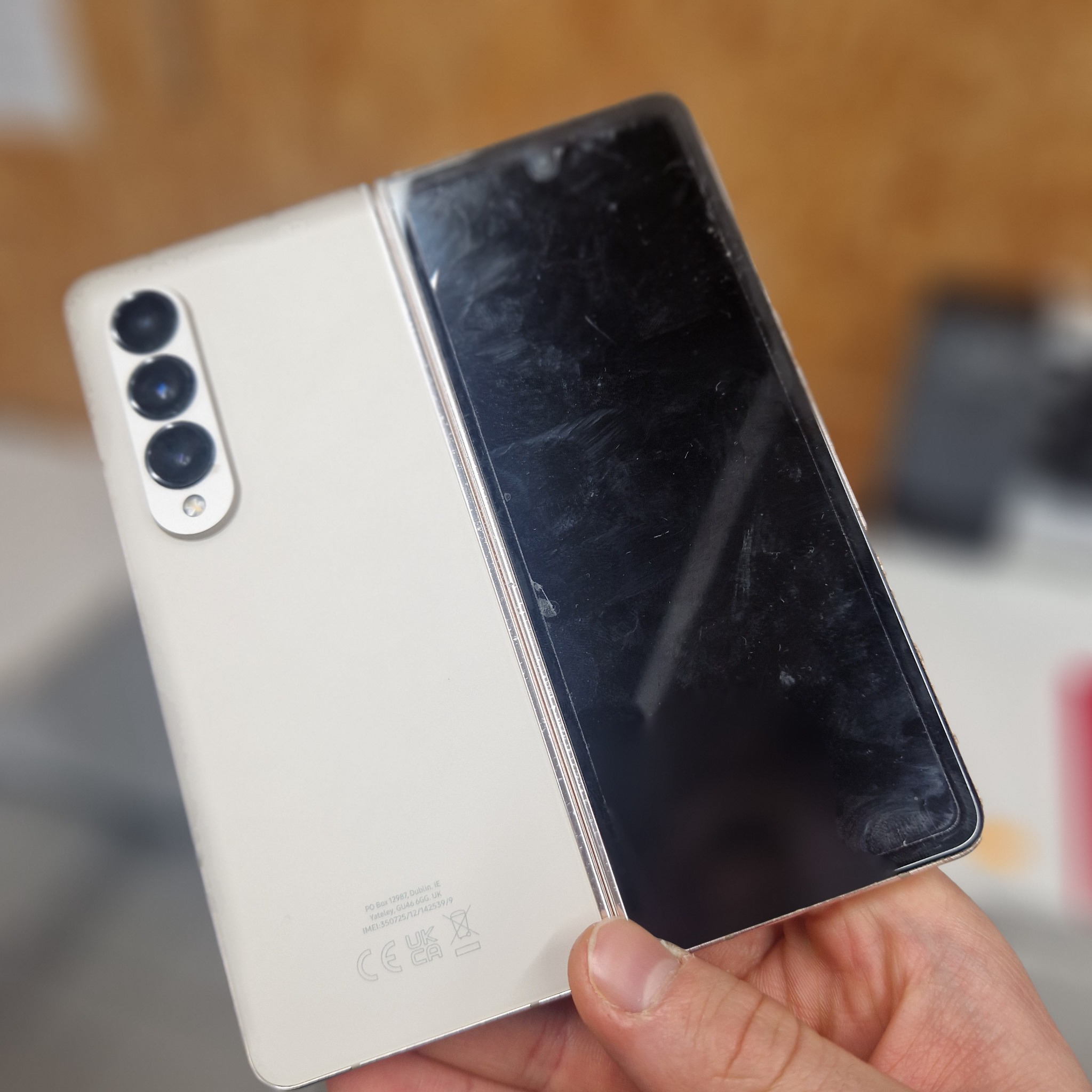 Rozłożony, składany smartfon w kolorze złamanej bieli trzymany w dłoni, ekran dotknięty smugami, widoczne trzy obiektywy aparatu na tylnej obudowie.