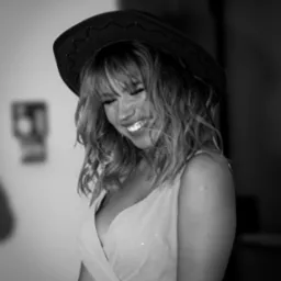 Portret uśmiechniętej kobiety w kowbojskim kapeluszu i sukience, czarno-białe zdjęcie z widocznym tatuażem na ramieniu.