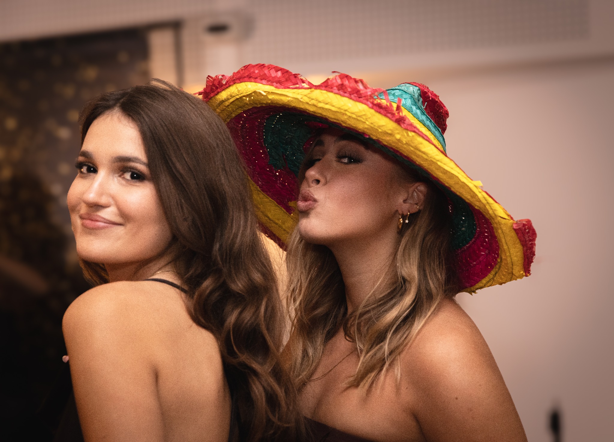 Dwie uśmiechnięte kobiety pozują do zdjęcia, jedna z nich w kolorowym, meksykańskim sombrero, robiąca dzióbek.