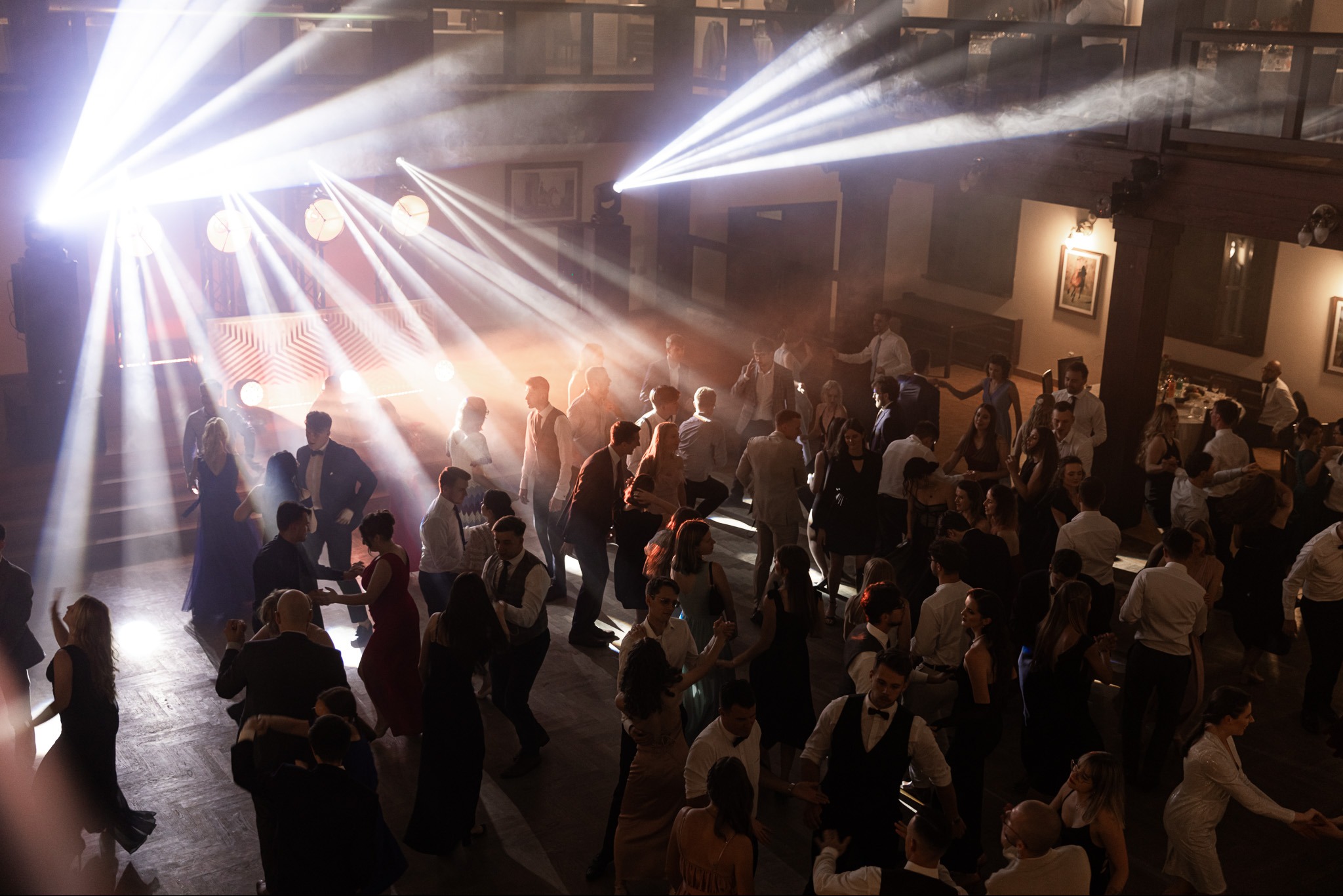 Parkiet taneczny wypełniony tańczącymi ludźmi podczas eventu, oświetlony reflektorami tworzącymi snopy światła, widoczne dekoracje i elementy architektoniczne sali.